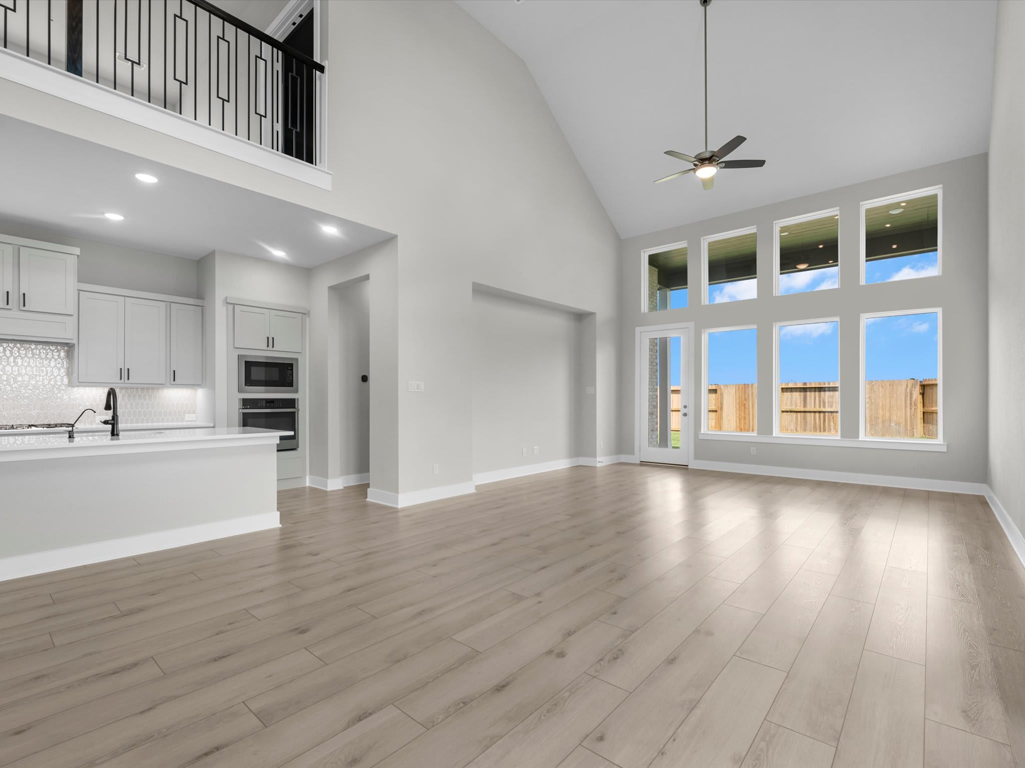 A spacious open-concept living area with light wood flooring leads toward large windows and a glass door in the background, with a modern white kitchen and an upstairs balcony railing in the foreground.