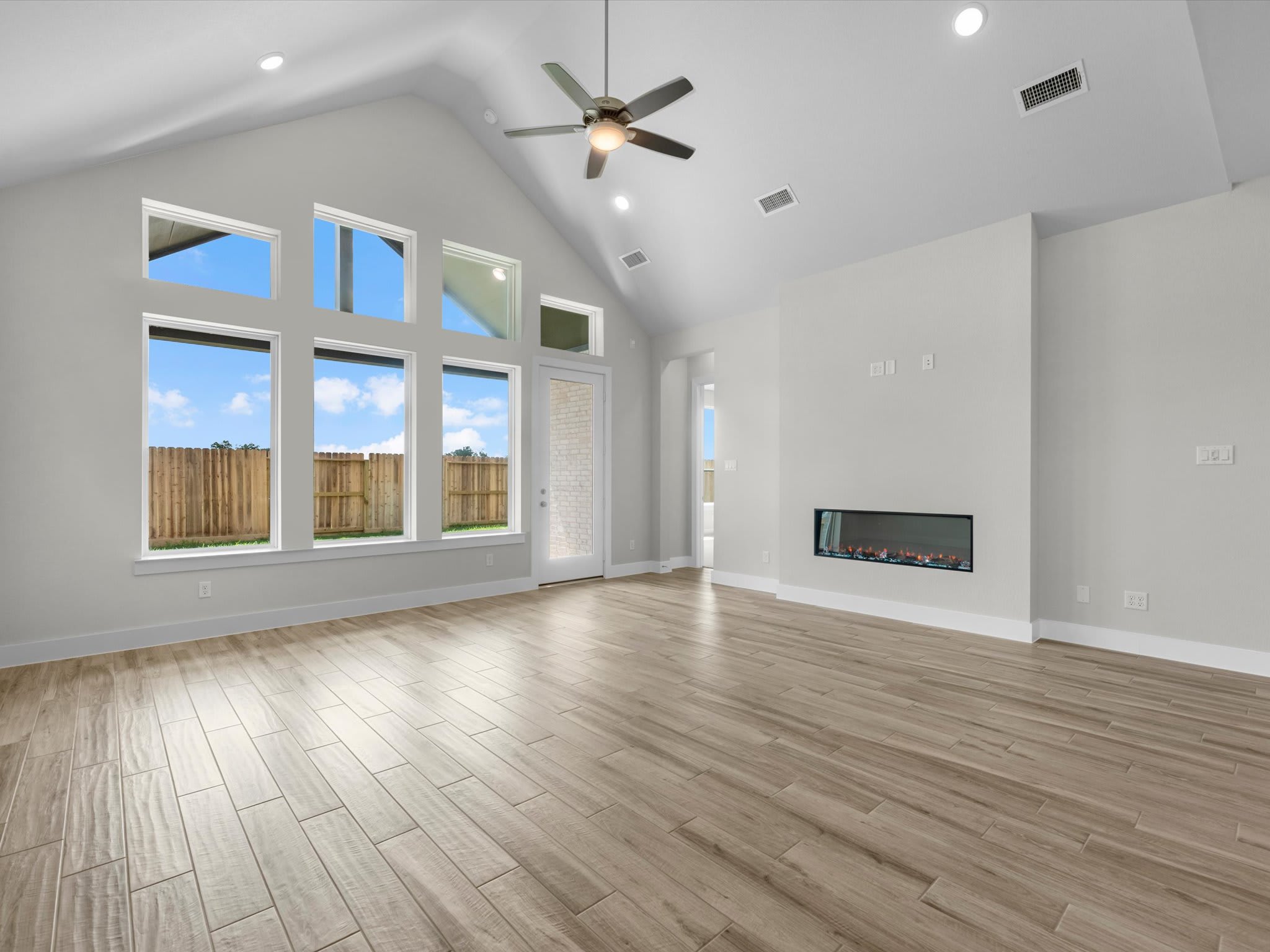 A spacious living room with light wood-plank flooring and a modern fireplace in the foreground opens up to large windows and a vaulted ceiling with a fan in the background.