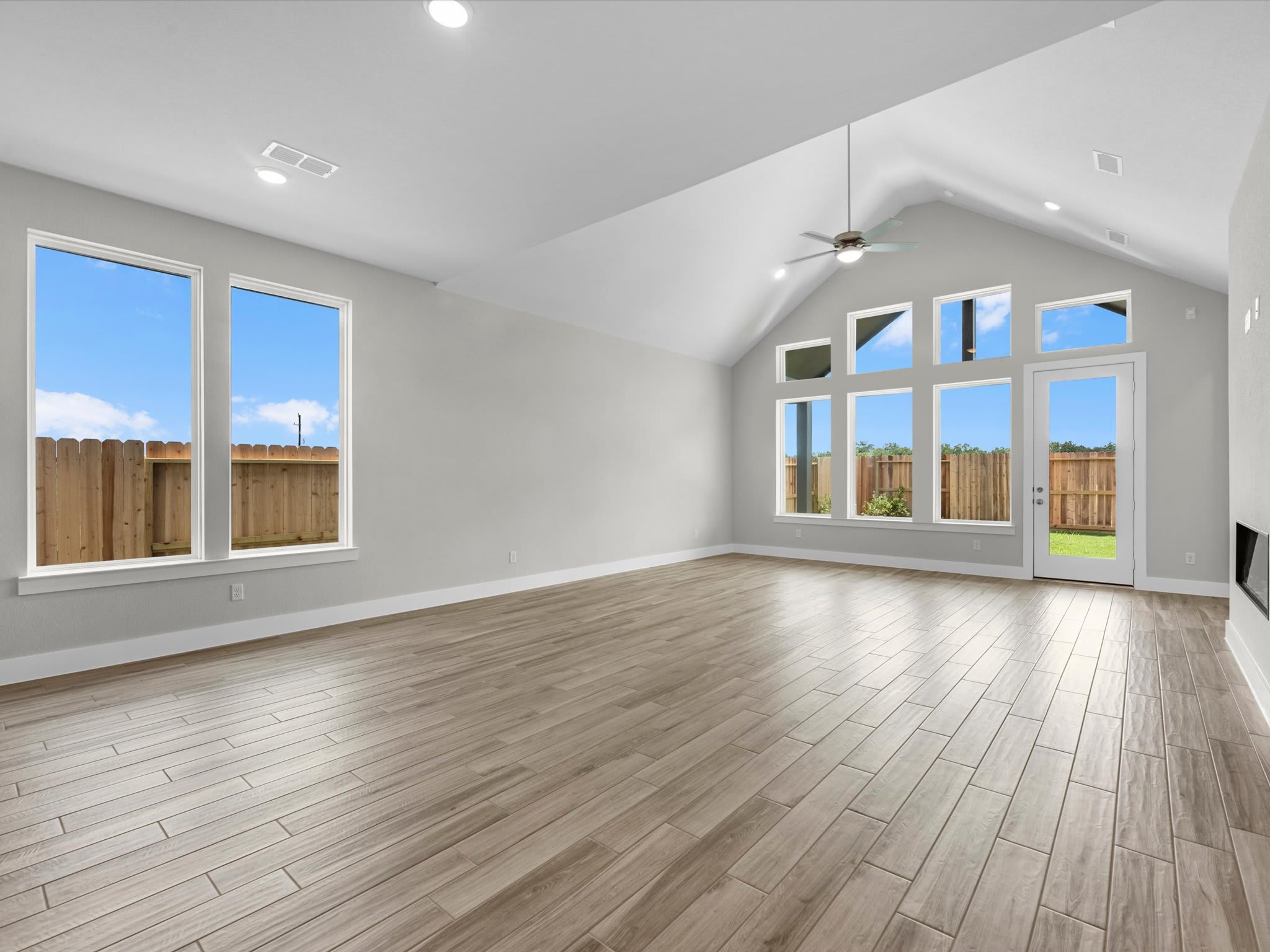 An empty room with light wood-plank flooring in the foreground and white walls with large windows overlooking a wooden fence in the background.