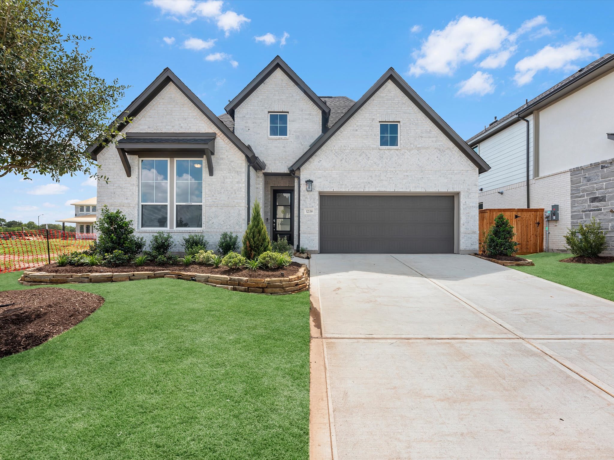 A modern white brick house with a grey garage door sits behind a manicured green lawn and concrete driveway under a bright blue sky.