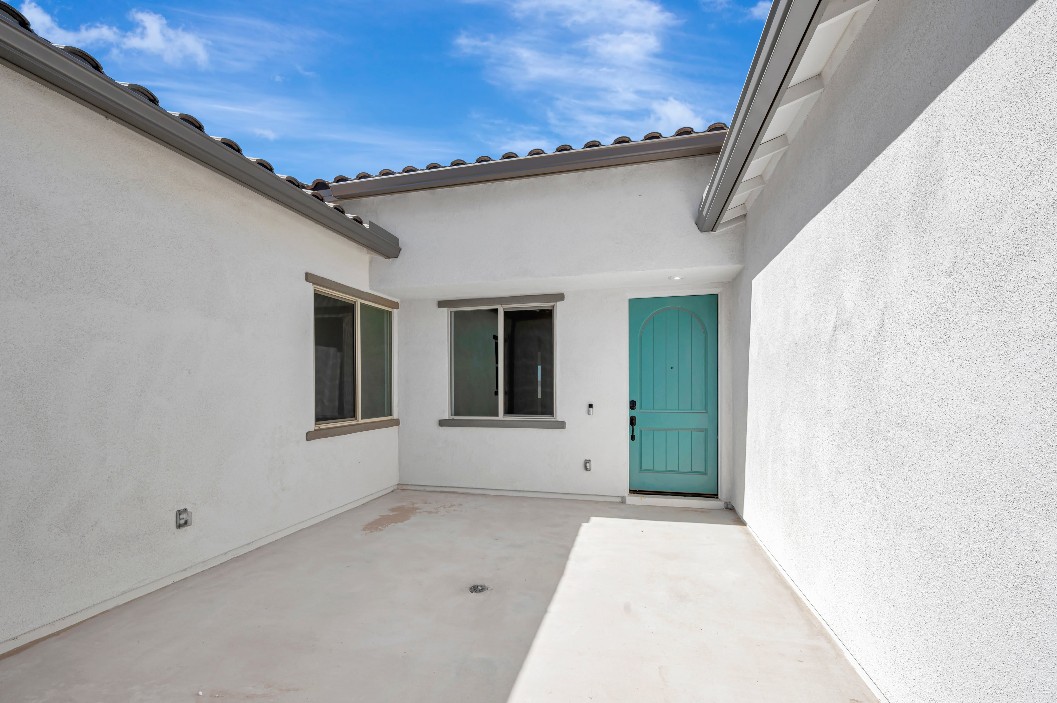 A light-colored concrete patio sits in the foreground, surrounded by white stucco walls and a turquoise door, under a bright blue sky with wispy clouds.