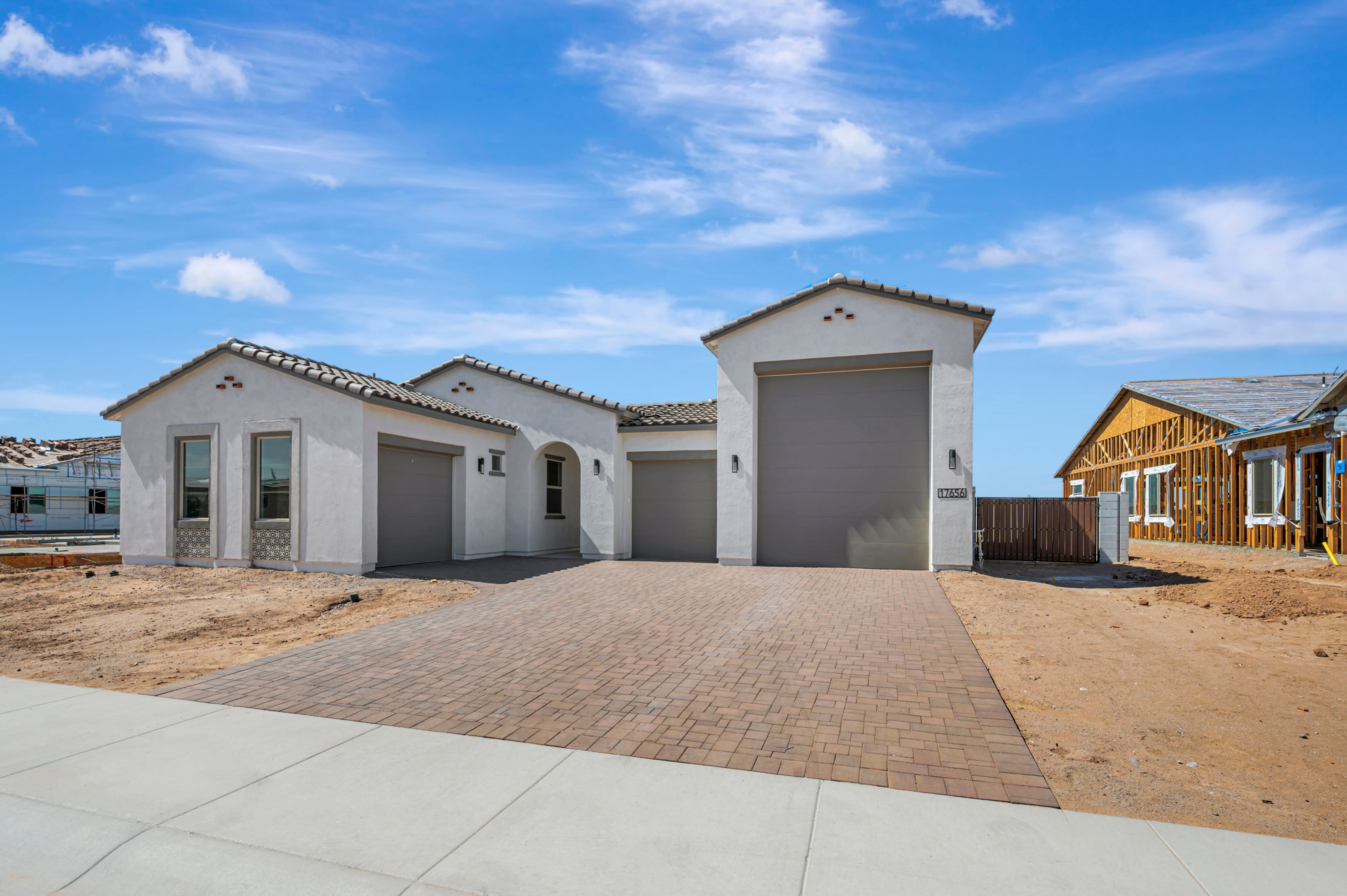 A paved driveway leads to a modern white house with multiple garages under a bright blue sky with wispy clouds, while a house under construction stands in the background.
