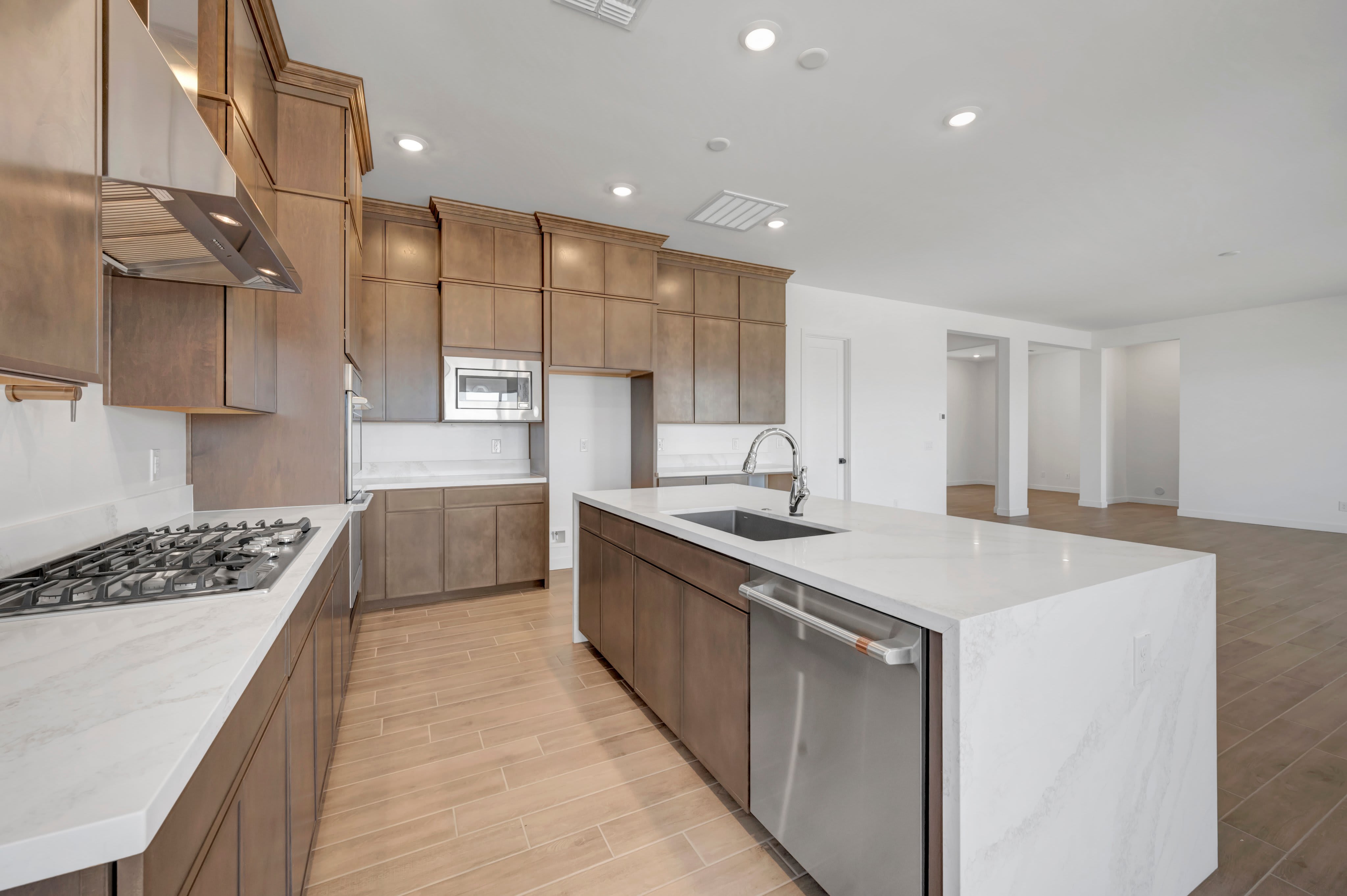 A modern kitchen features a white quartz island with a built-in sink and dishwasher in the foreground, set against a background of brown wooden cabinetry and light-colored tile flooring.