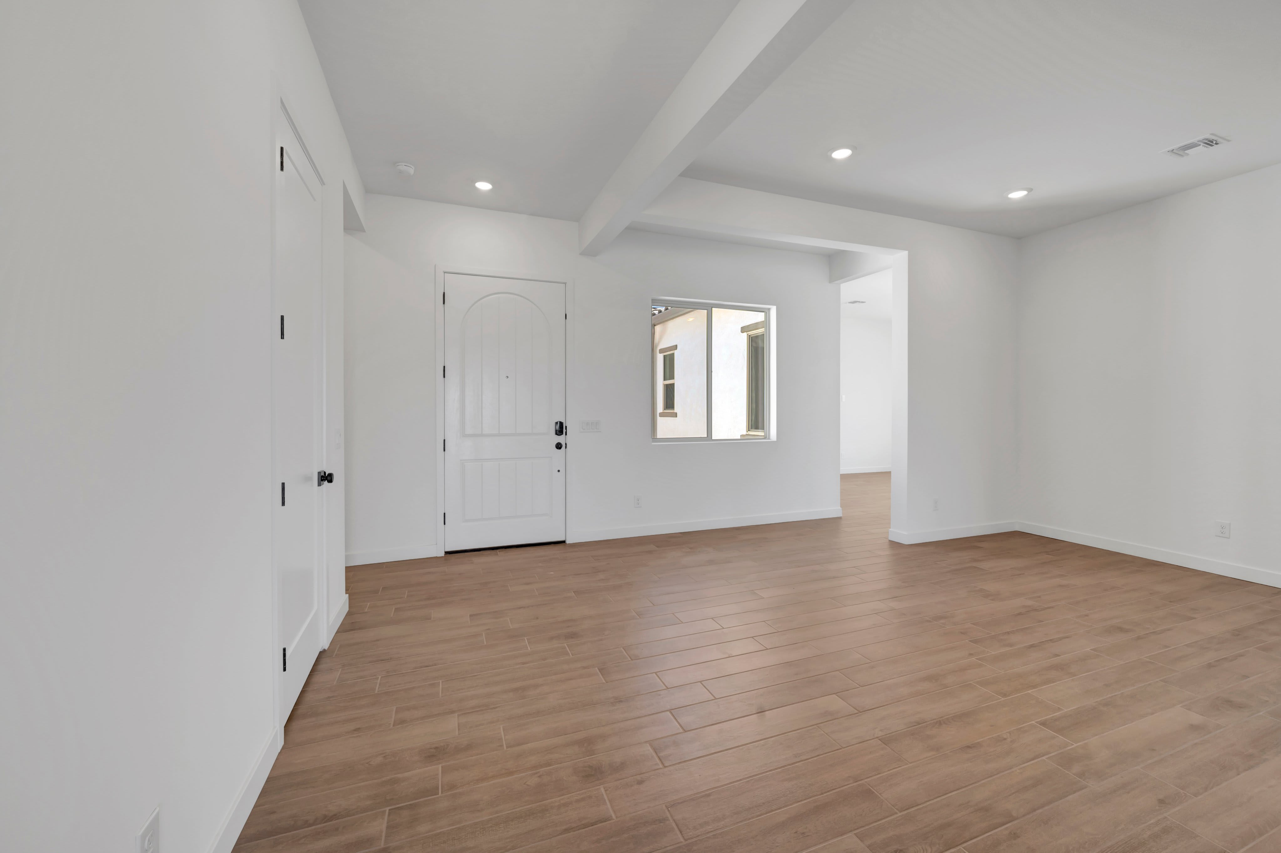 Light-colored wood flooring fills the foreground of an empty white room featuring a closed door and a window in the background.