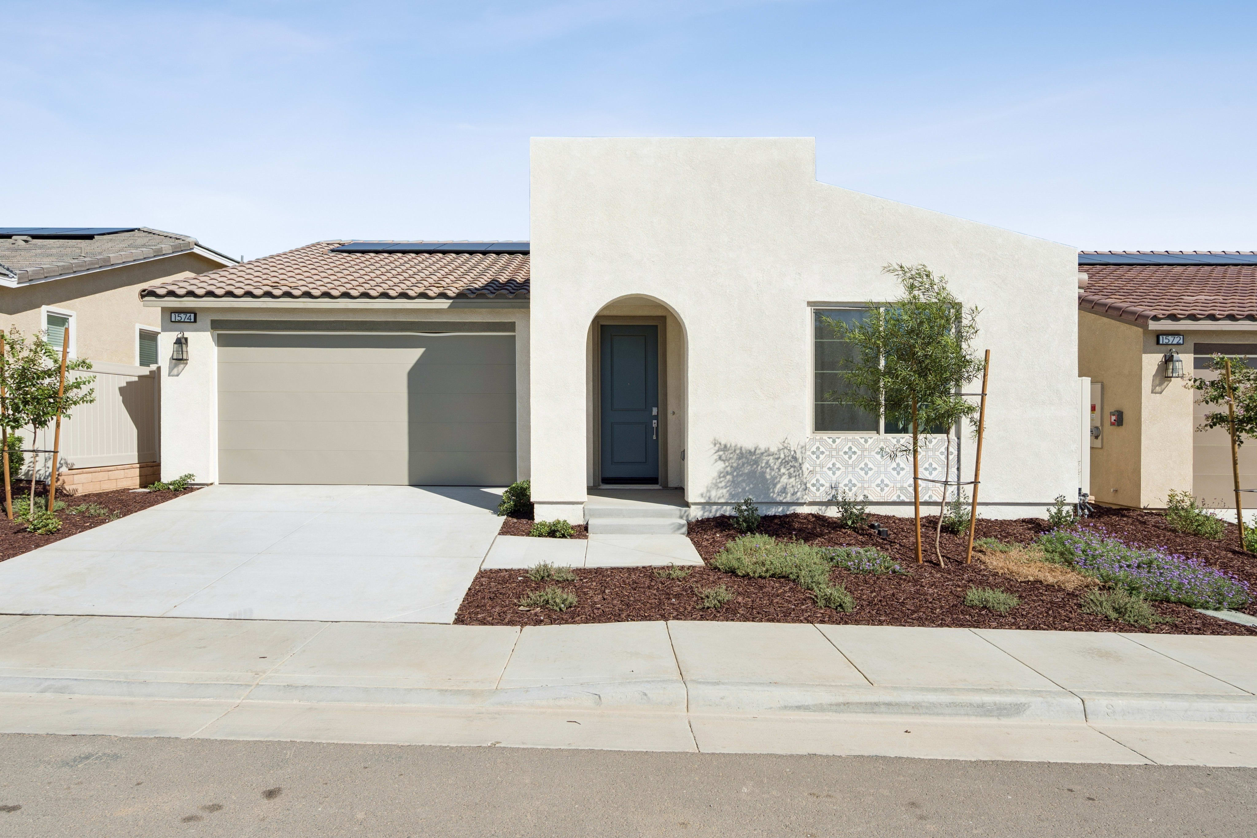 A modern white stucco house with a grey garage door sits behind a concrete driveway and landscaped garden under a clear blue sky.