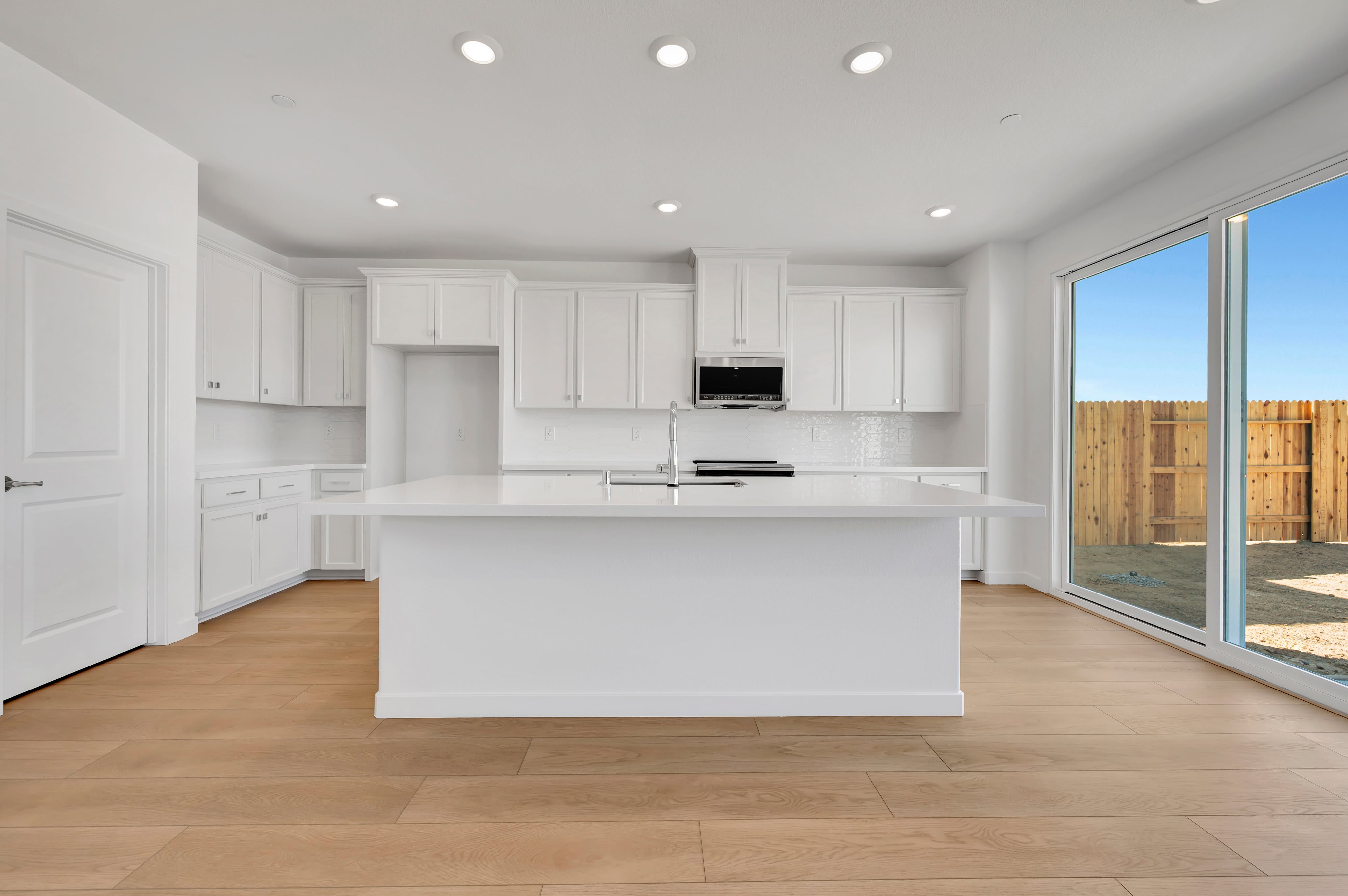 A white kitchen island sits on light wood floors in the foreground, with white cabinetry and a sliding glass door leading to a fenced yard in the background.