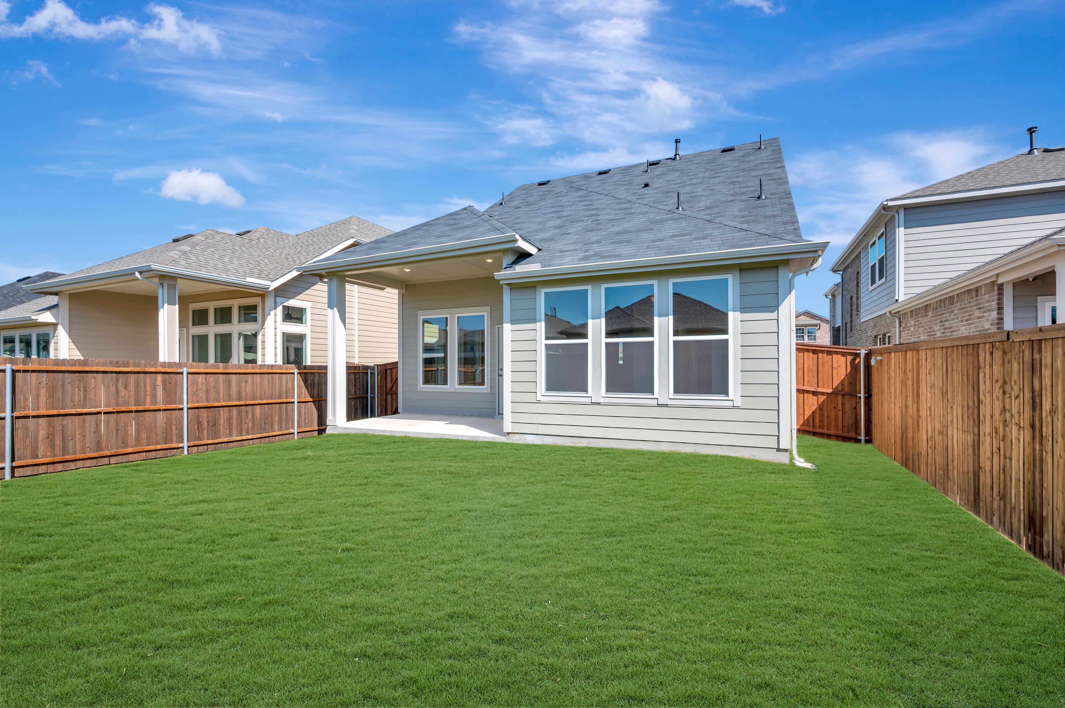A lush green lawn fills the foreground, leading to a modern grey house and wooden fences under a bright blue sky.