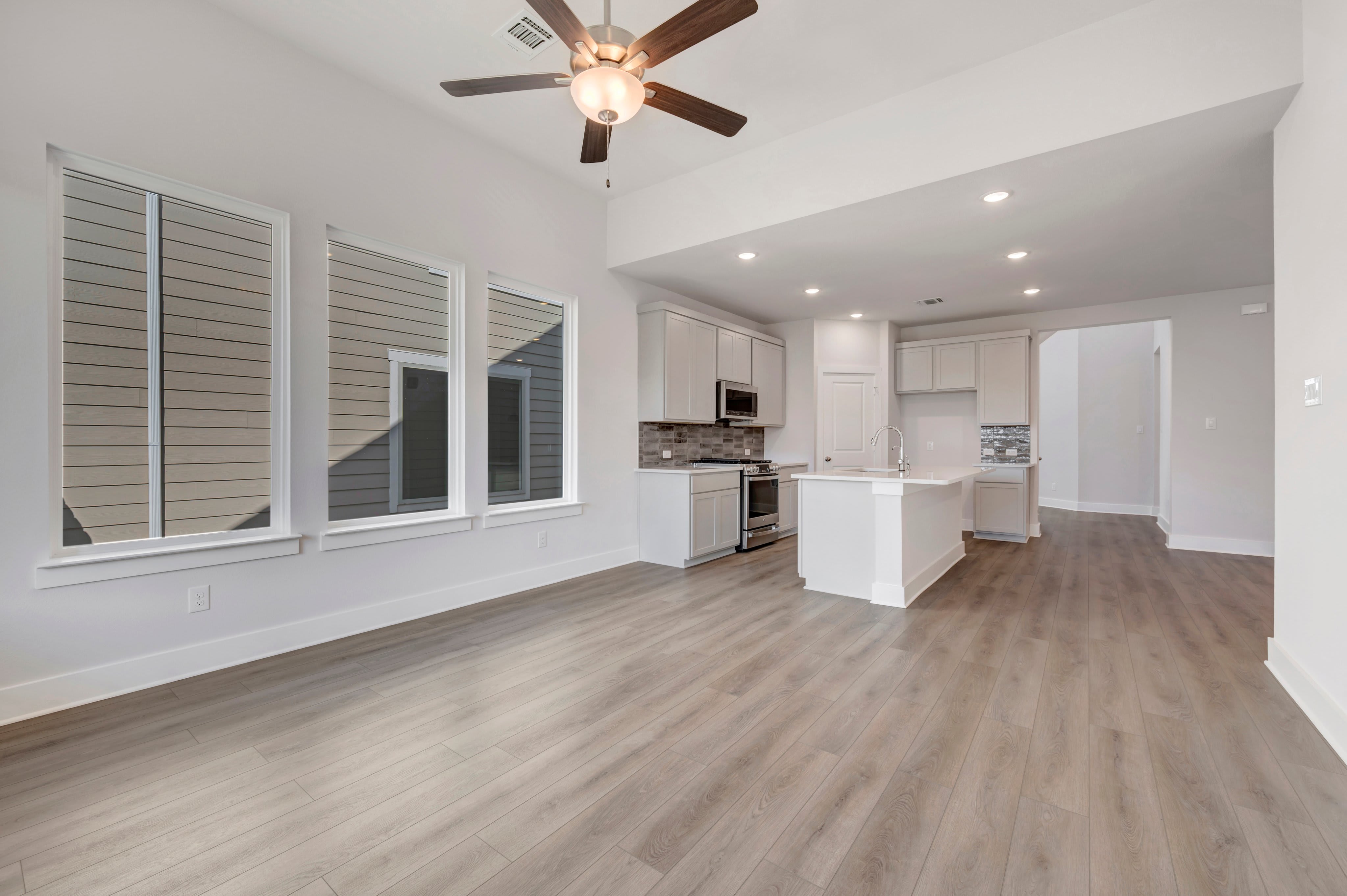 An empty room with light wood flooring and large windows in the foreground opens into a white modern kitchen in the background.