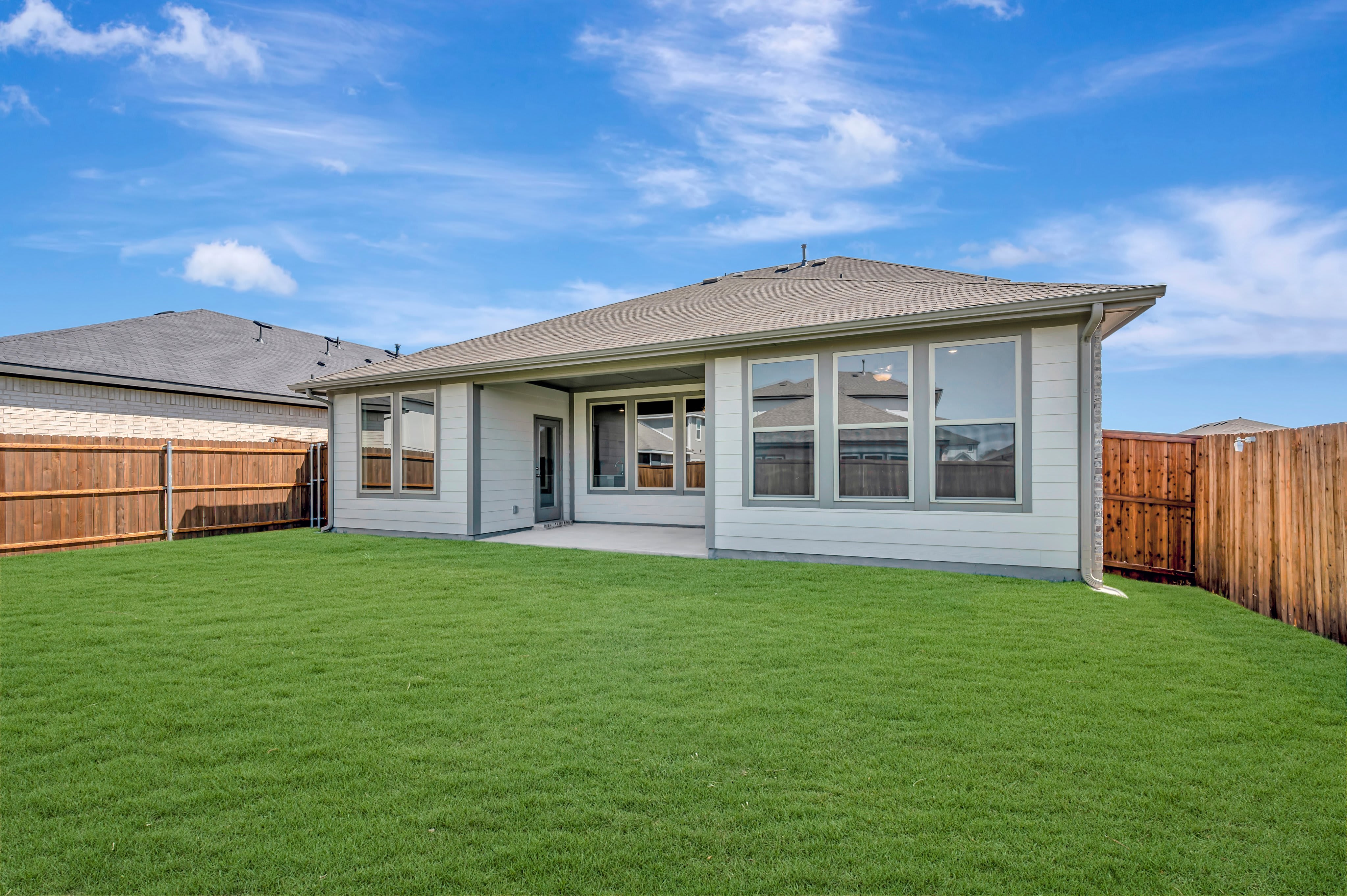 A lush green lawn fills the foreground, leading to a light-colored house with large windows set against a bright blue sky with wispy clouds.