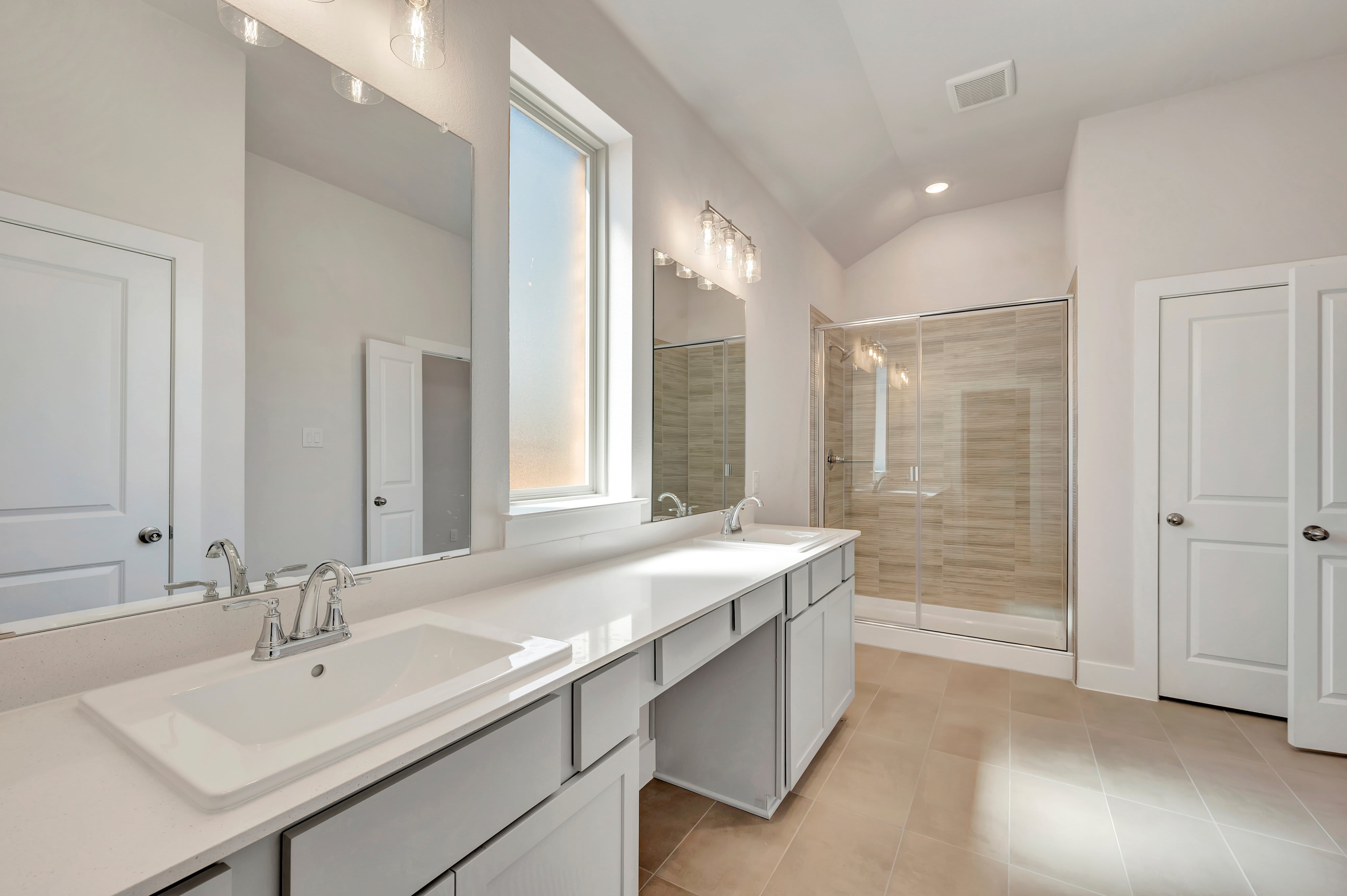 A white double-sink vanity with mirrors sits in the foreground of a bright bathroom featuring a glass-enclosed shower and white doors in the background.