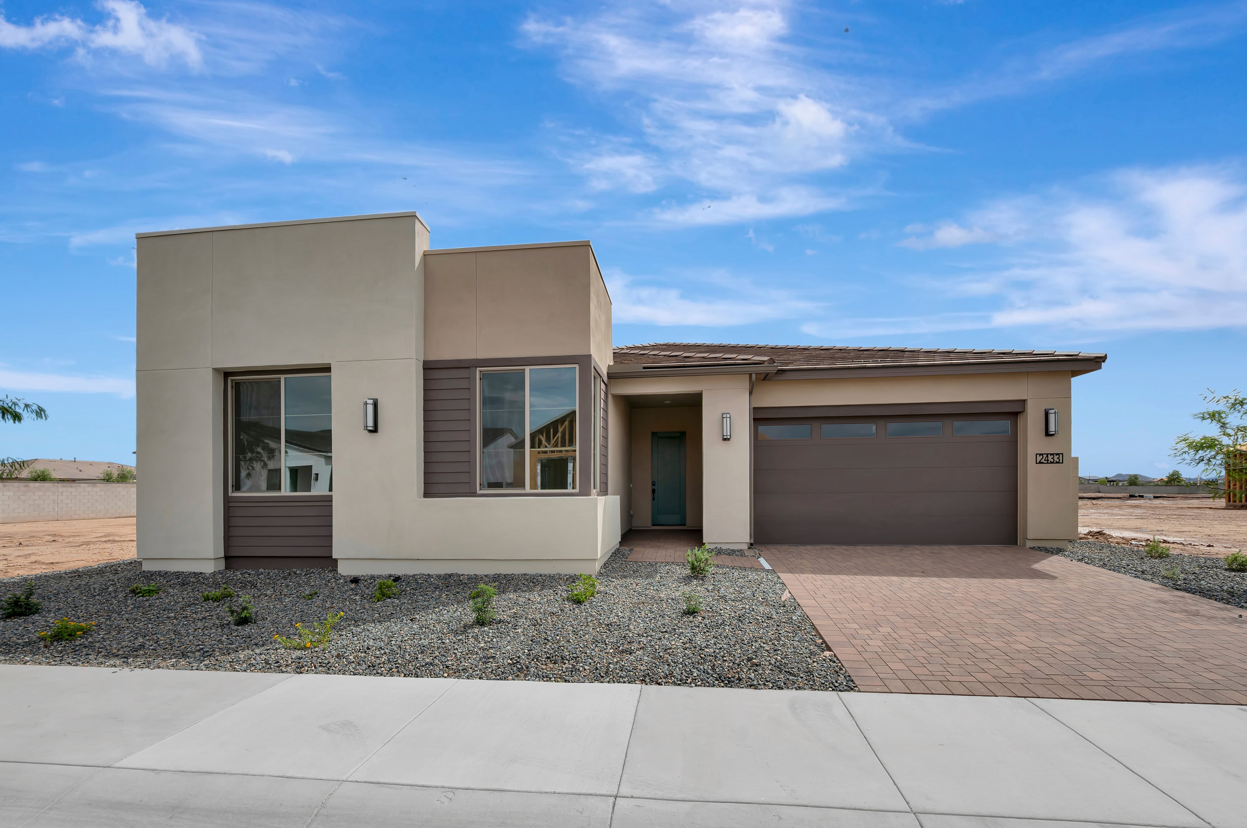 A modern single-story home with a brown garage door sits behind a gravel landscape and paved driveway under a bright blue sky with wispy clouds.