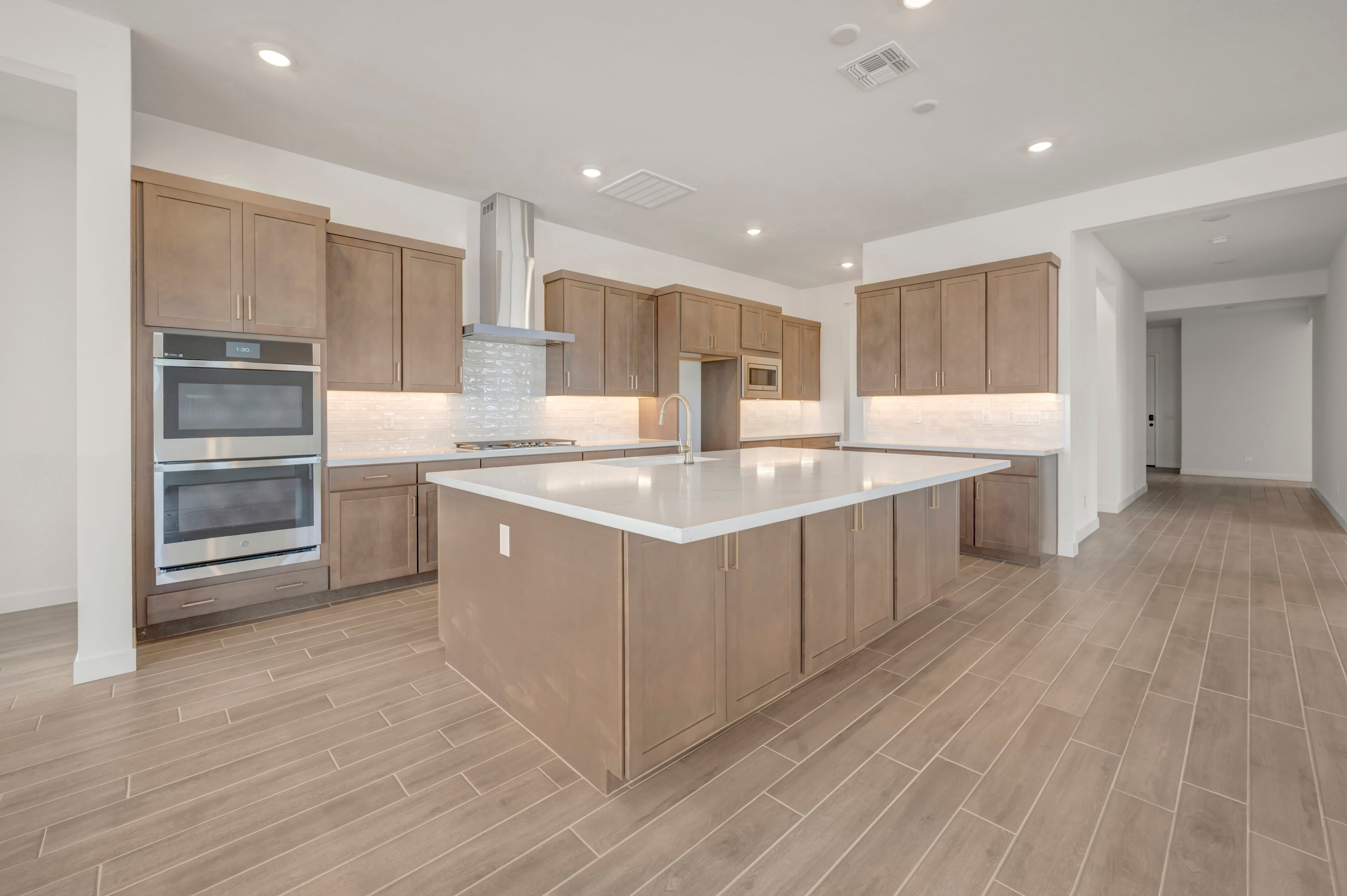 A large kitchen island with a white countertop sits in the foreground of a modern kitchen featuring light wood cabinetry and stainless steel appliances.
