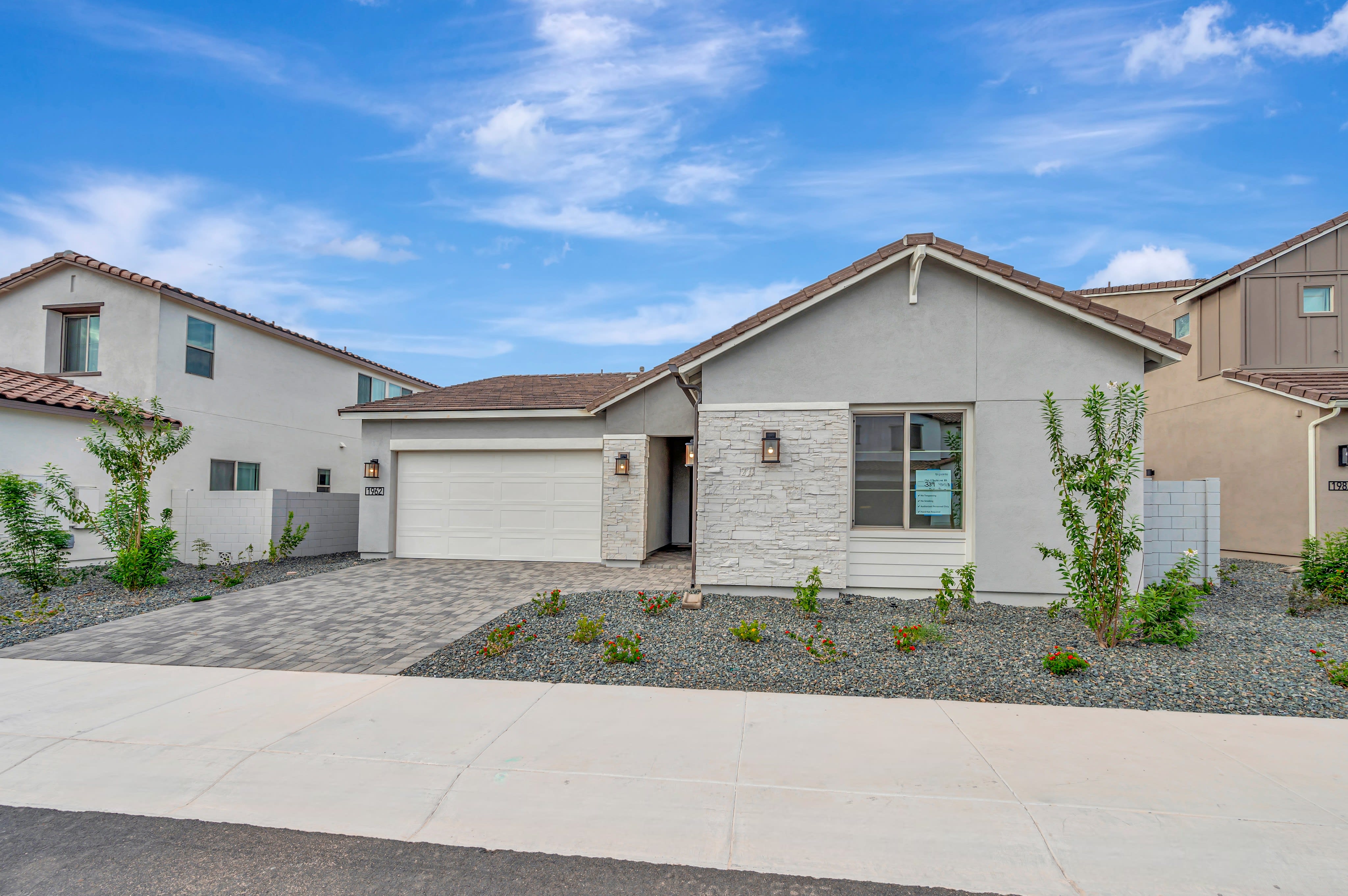 A modern single-story home with a grey exterior and stone accents sits behind a concrete sidewalk and gravel landscaping under a bright blue sky.