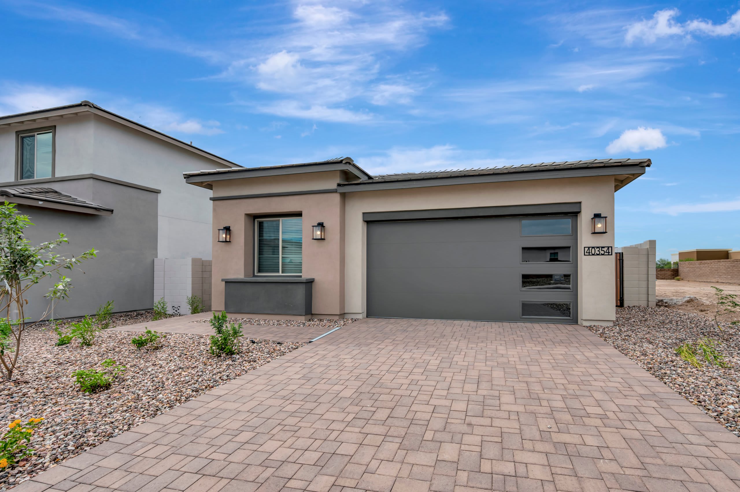 A paved driveway leads to a modern grey house with a large garage door under a bright blue sky with wispy clouds.