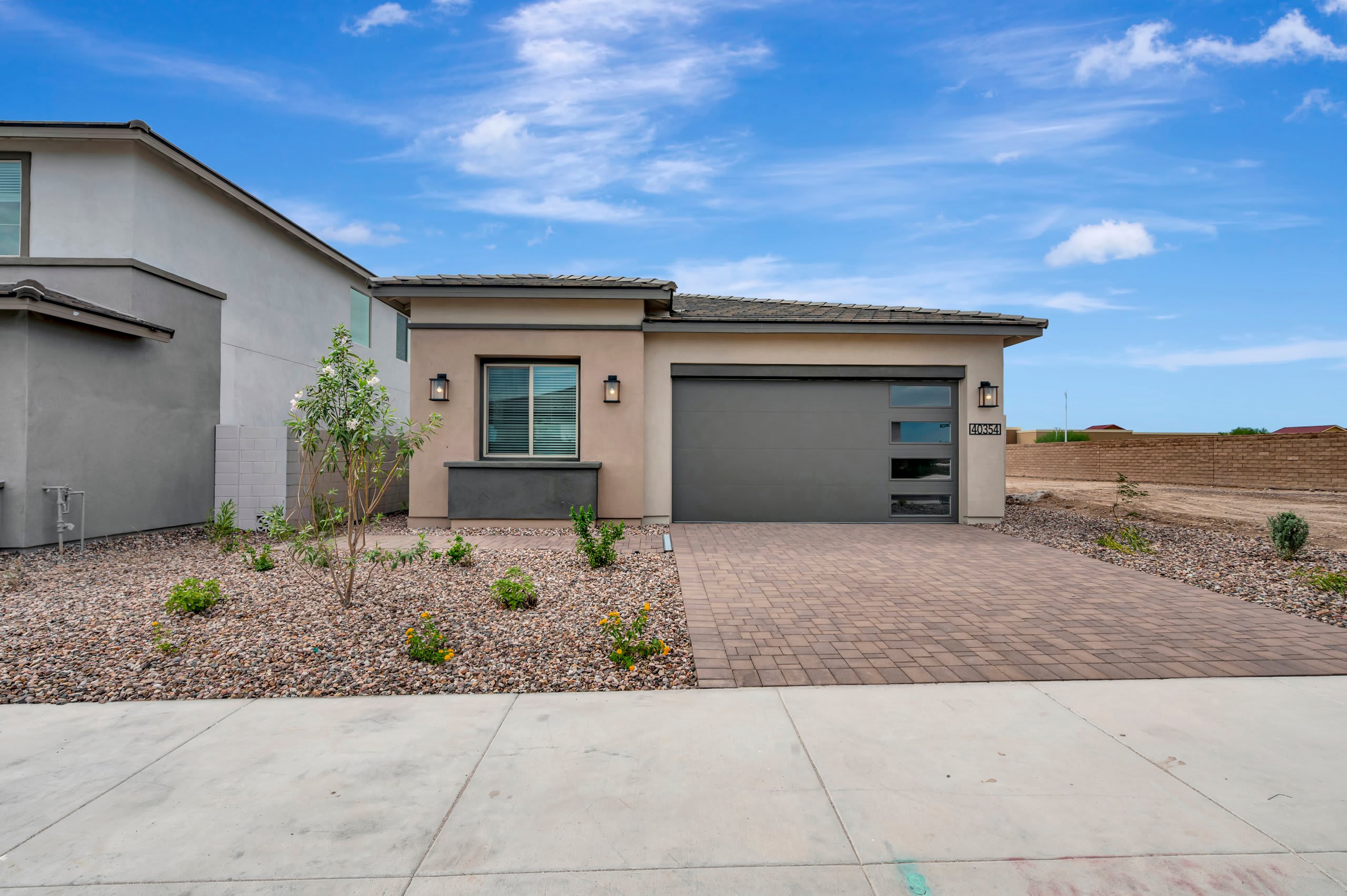 A modern grey house with a paved driveway and gravel landscaping sits under a bright blue sky with wispy clouds.