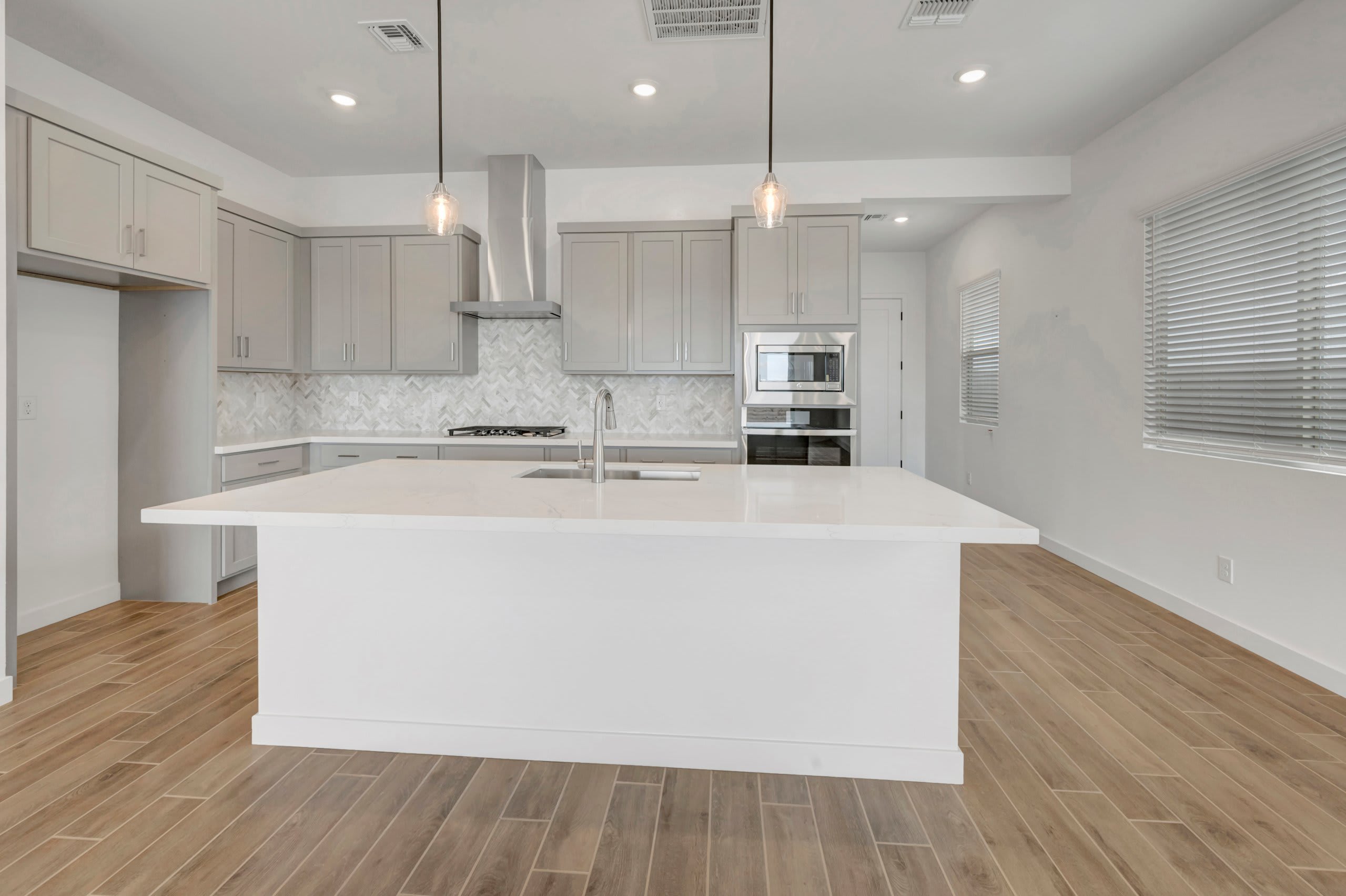 A large white kitchen island sits in the foreground of a modern kitchen featuring grey cabinetry, stainless steel appliances, and light wood flooring.