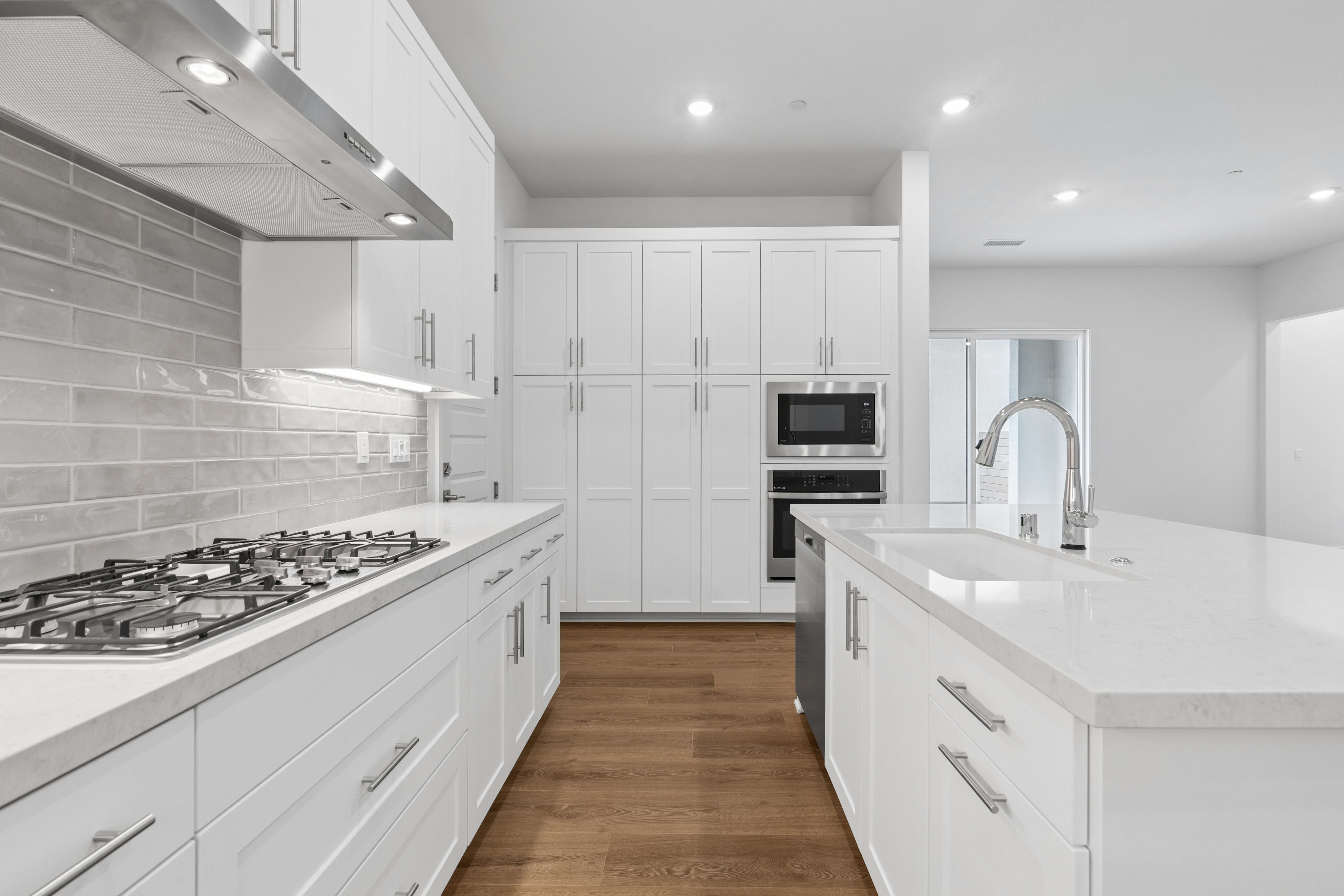 A modern white kitchen features a gas stovetop and white countertops in the foreground, with white cabinetry and built-in appliances against a light-colored wall in the background.