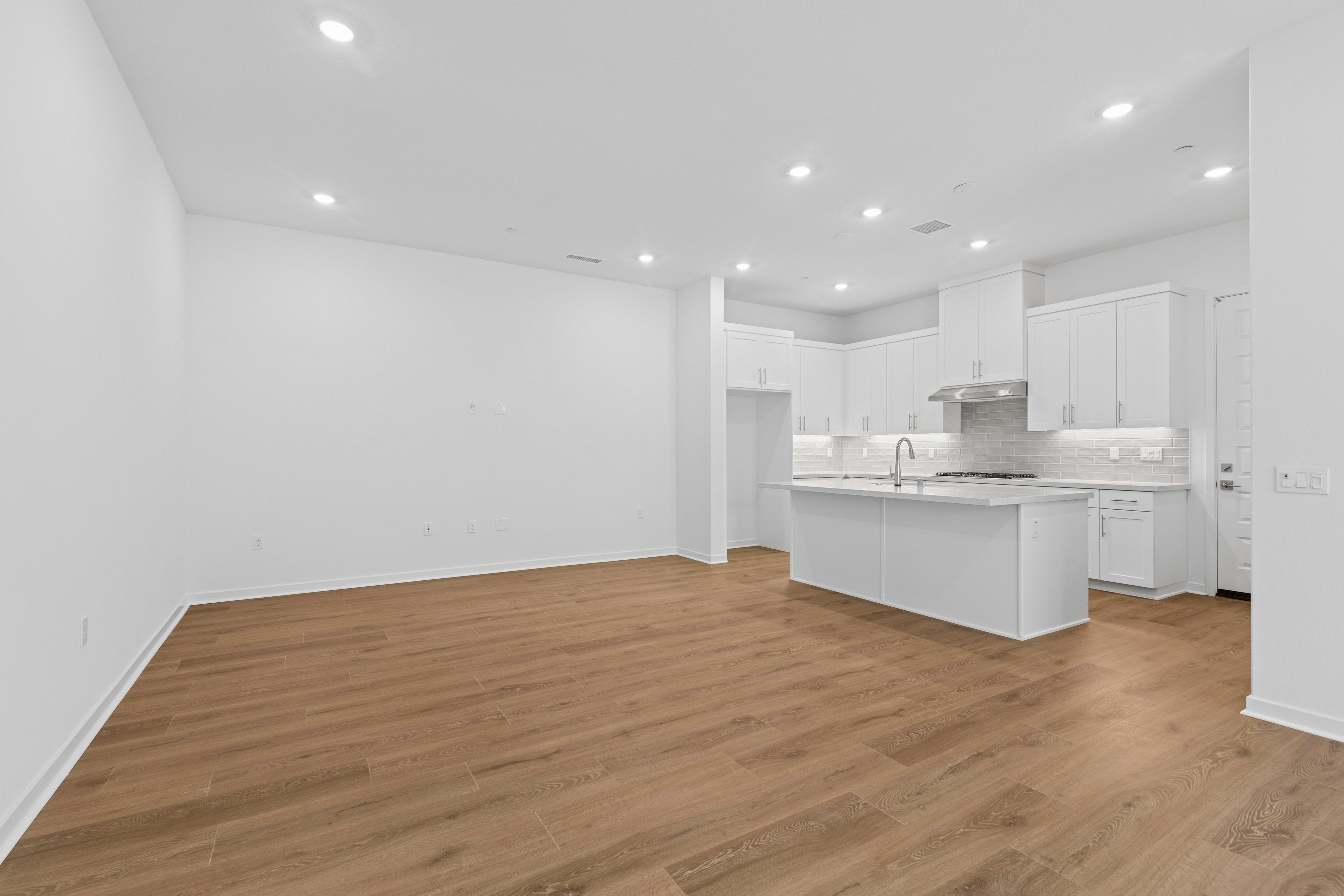 An empty room with light brown wood flooring leads into a modern white kitchen in the background.