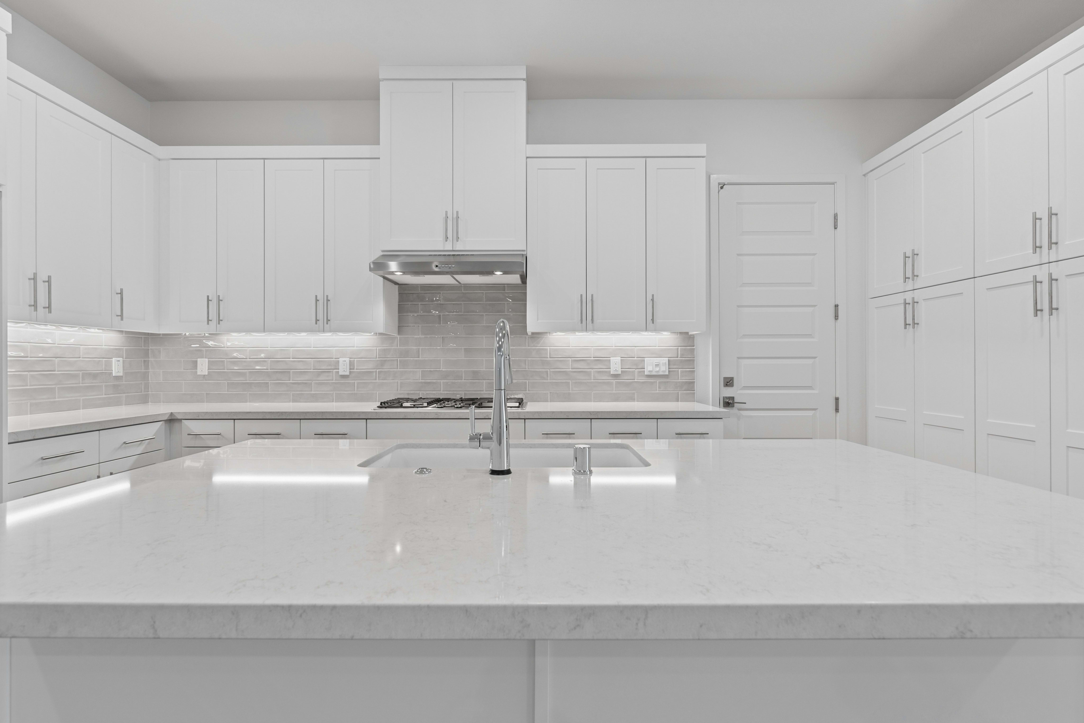 A white quartz kitchen island with a sink sits in the foreground of a modern kitchen featuring white cabinetry and a tiled backsplash.