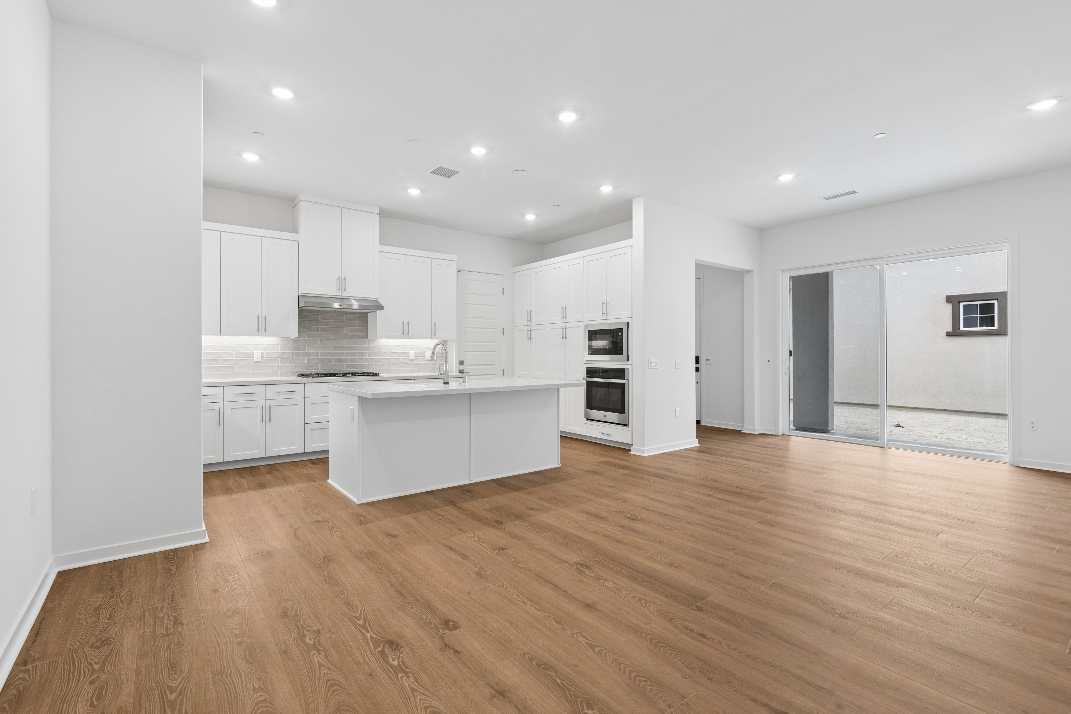 A modern white kitchen with a central island sits in the background of a spacious room with light wood flooring in the foreground.