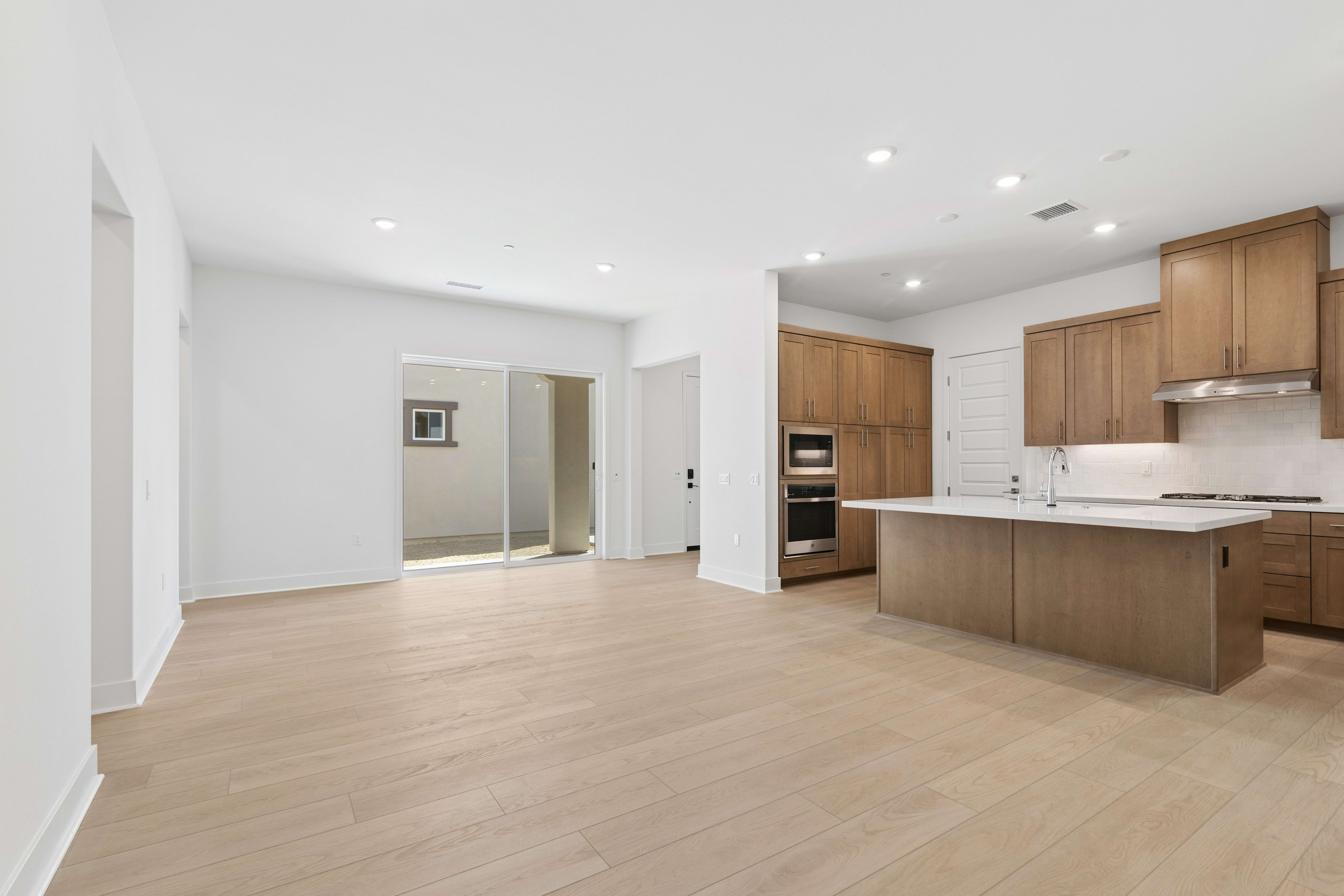 An open-concept living area with light wood flooring in the foreground leads to a modern kitchen with wooden cabinetry and a sliding glass door in the background.