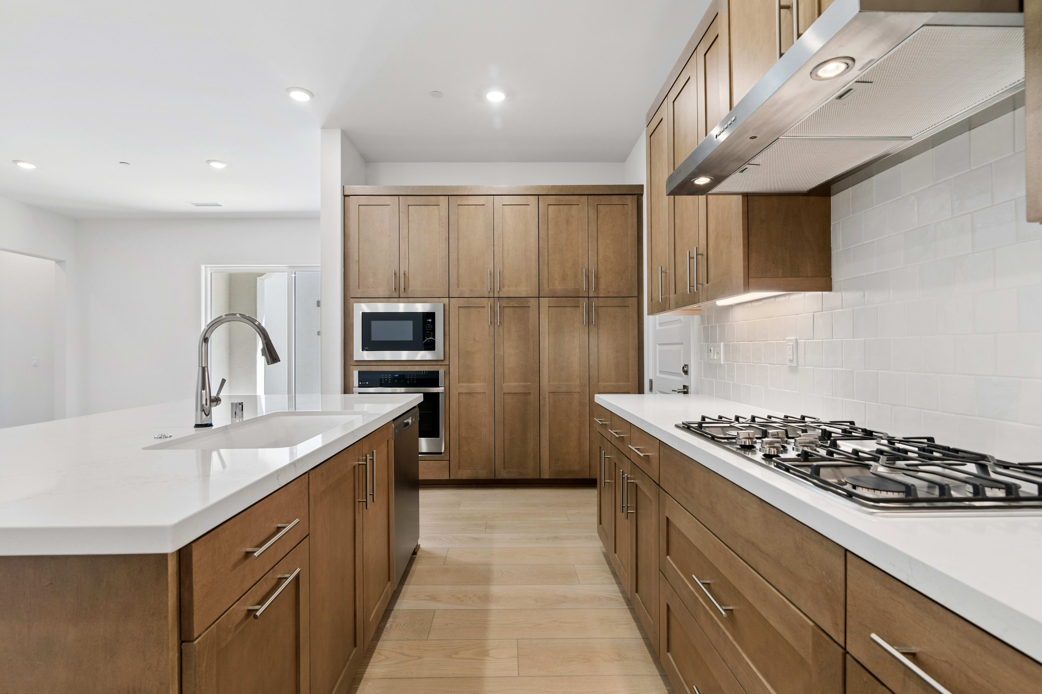 A modern kitchen features a white quartz island with a sink in the foreground and light wood cabinetry with built-in appliances in the background.