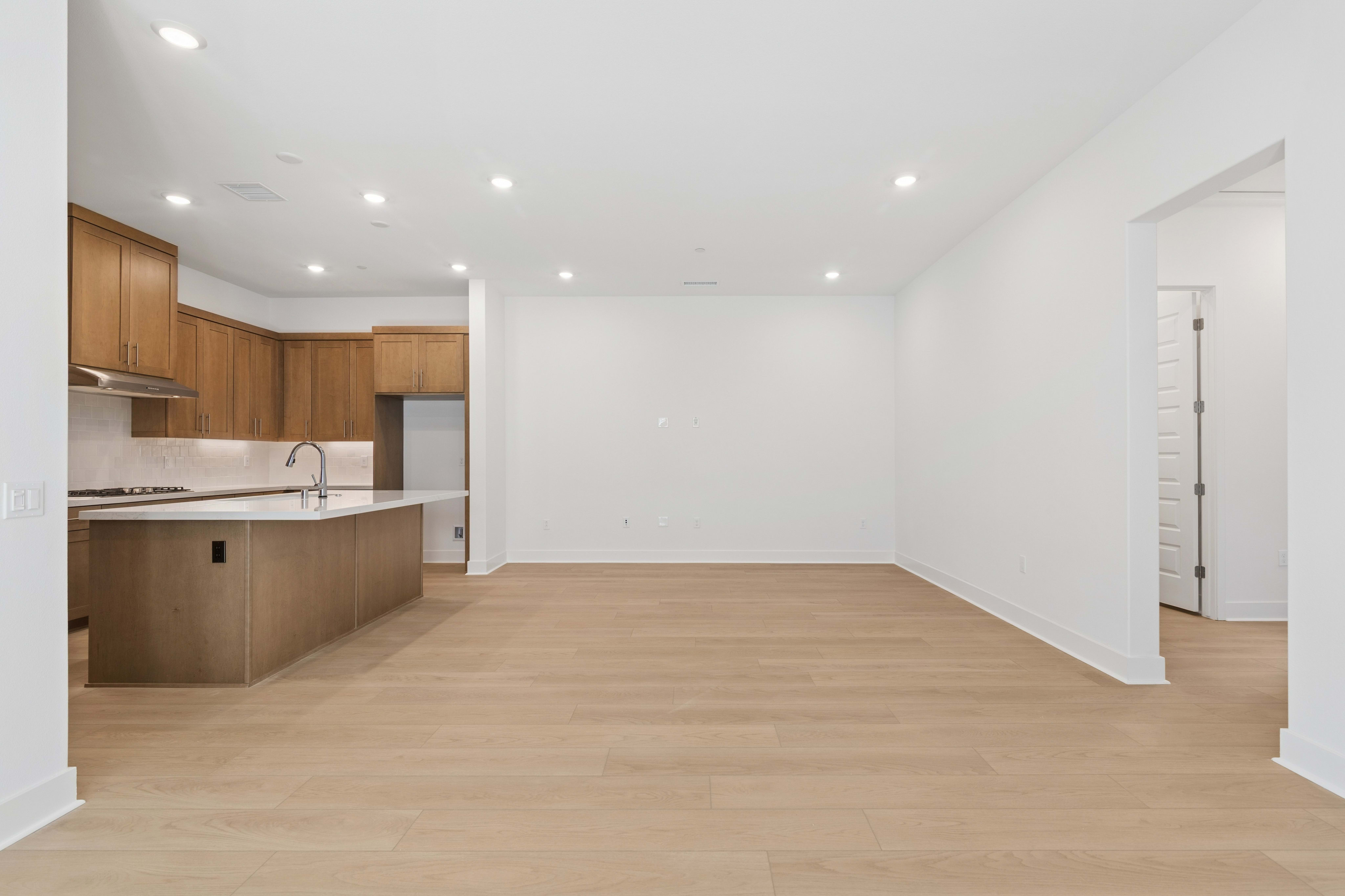 A modern open-concept living space features a kitchen with light wood cabinetry and white countertops in the foreground, opening into a large empty room with light hardwood floors and white walls in the background.