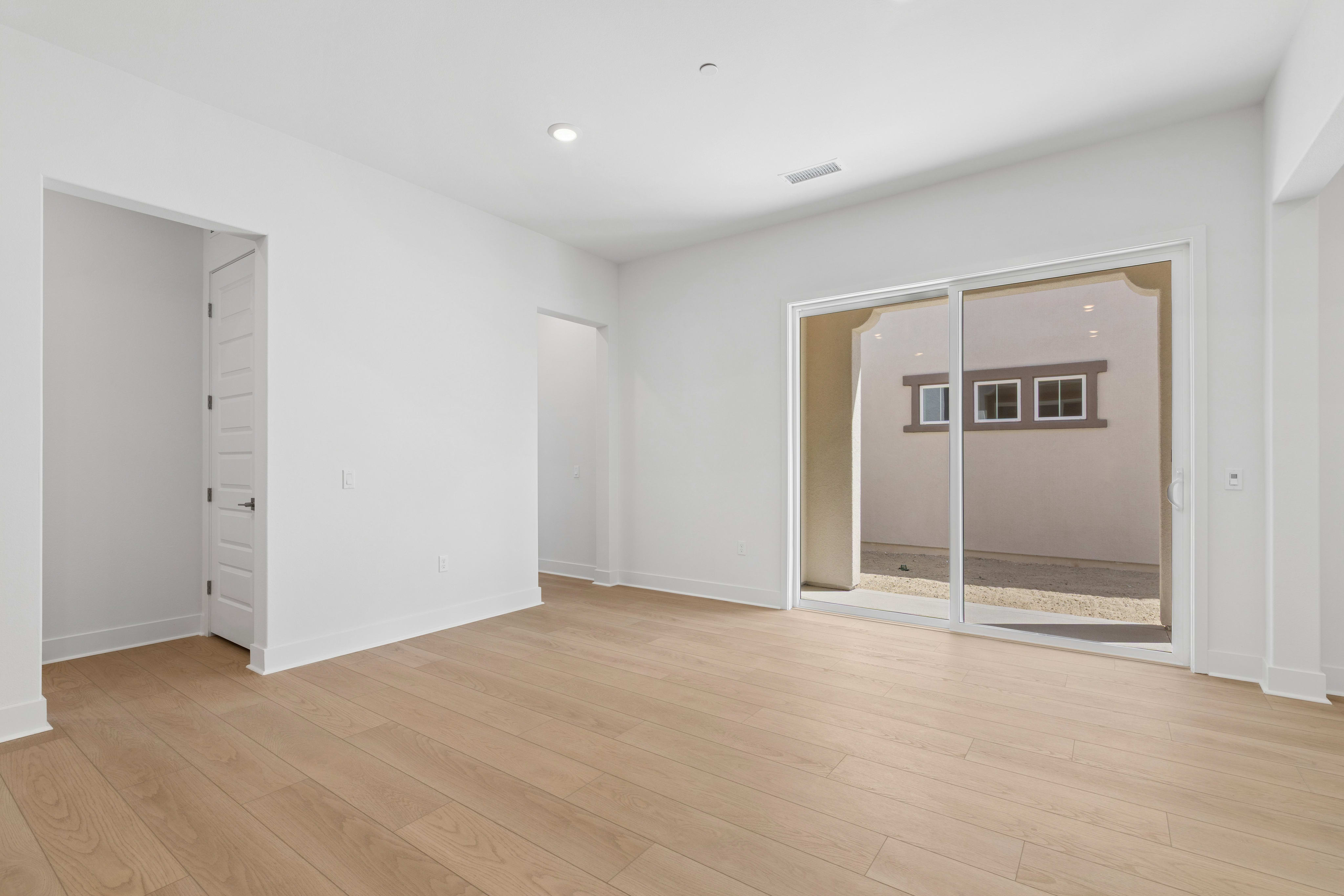 An empty room with light wood flooring and white walls features a sliding glass door leading to an outdoor patio area.