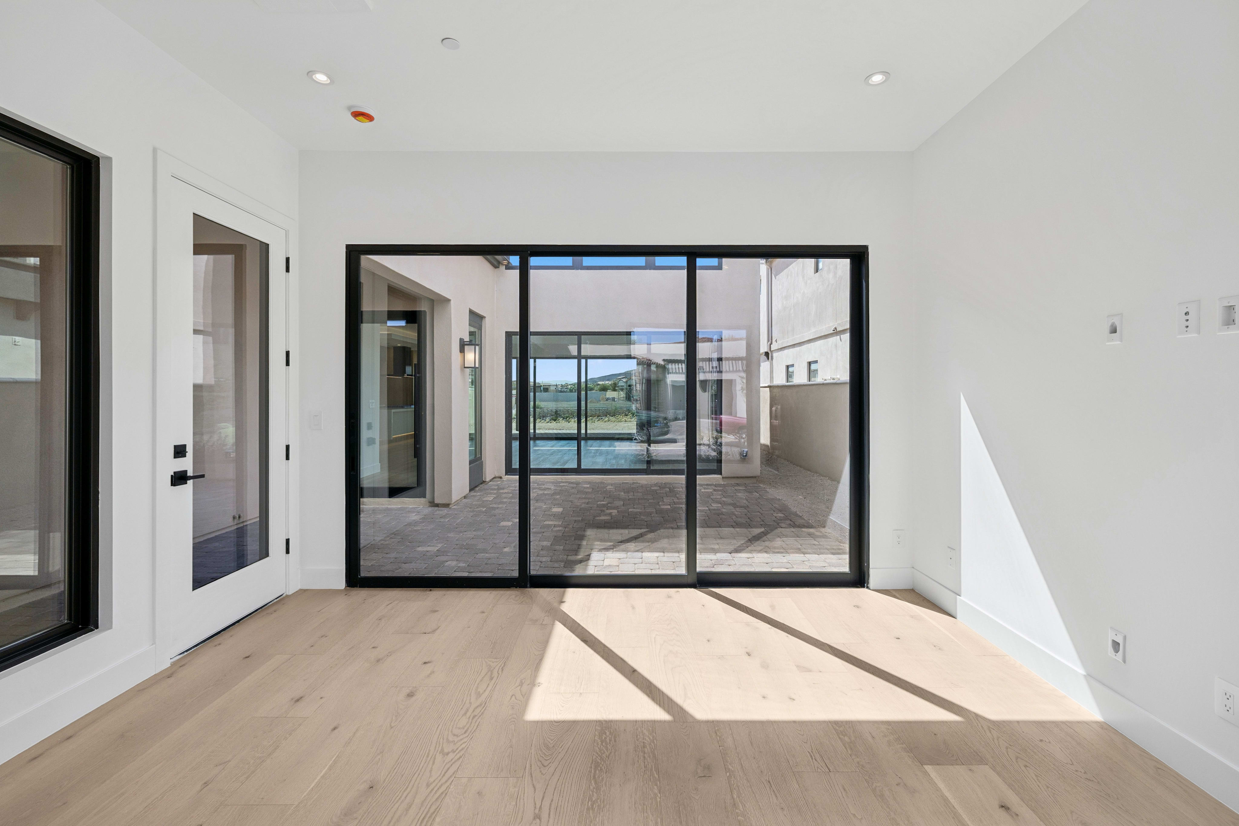 An empty room with light wood floors and white walls opens through large sliding glass doors to a paved outdoor patio.