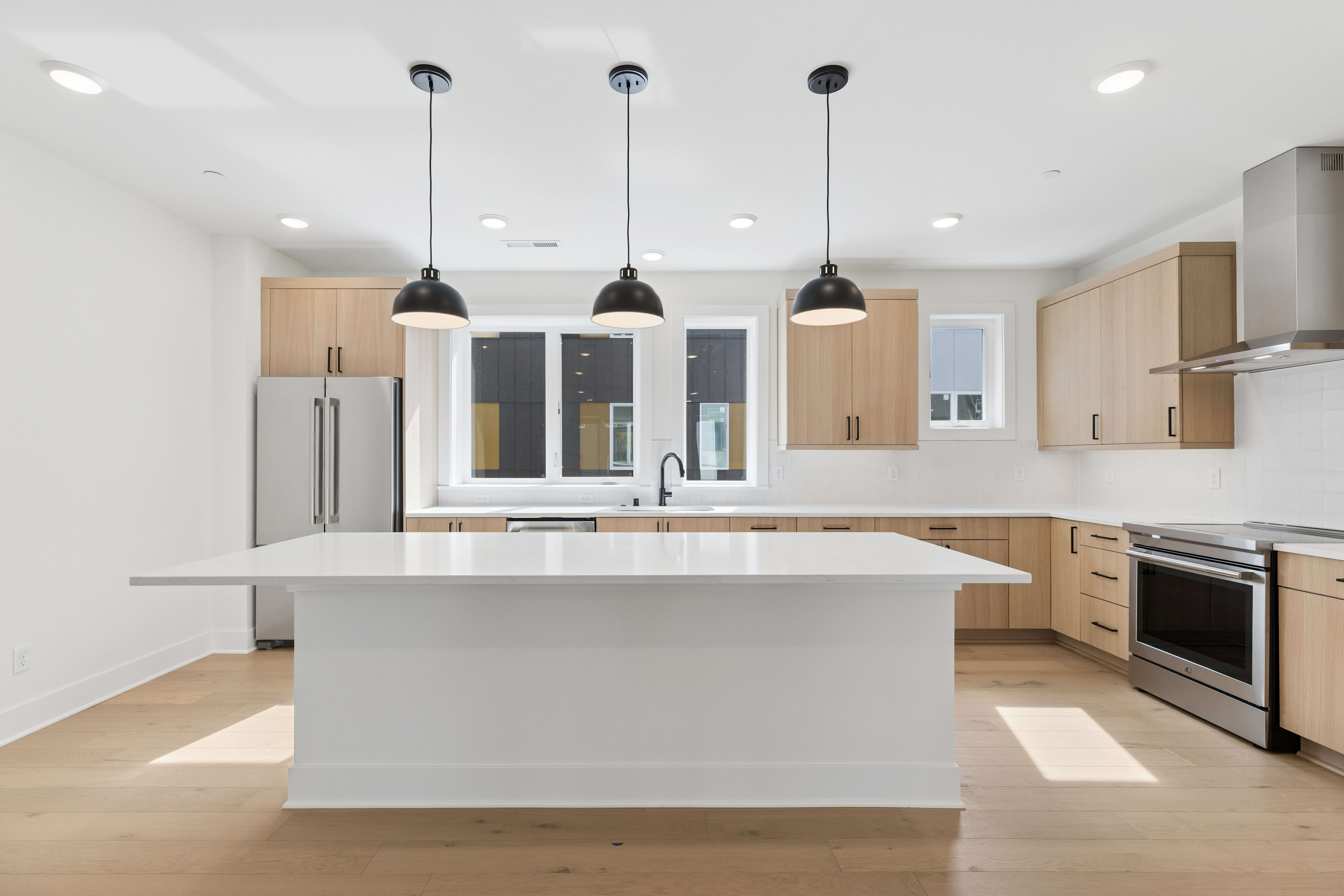 A white kitchen island sits in the foreground of a modern kitchen featuring light wood cabinets and three black pendant lights.