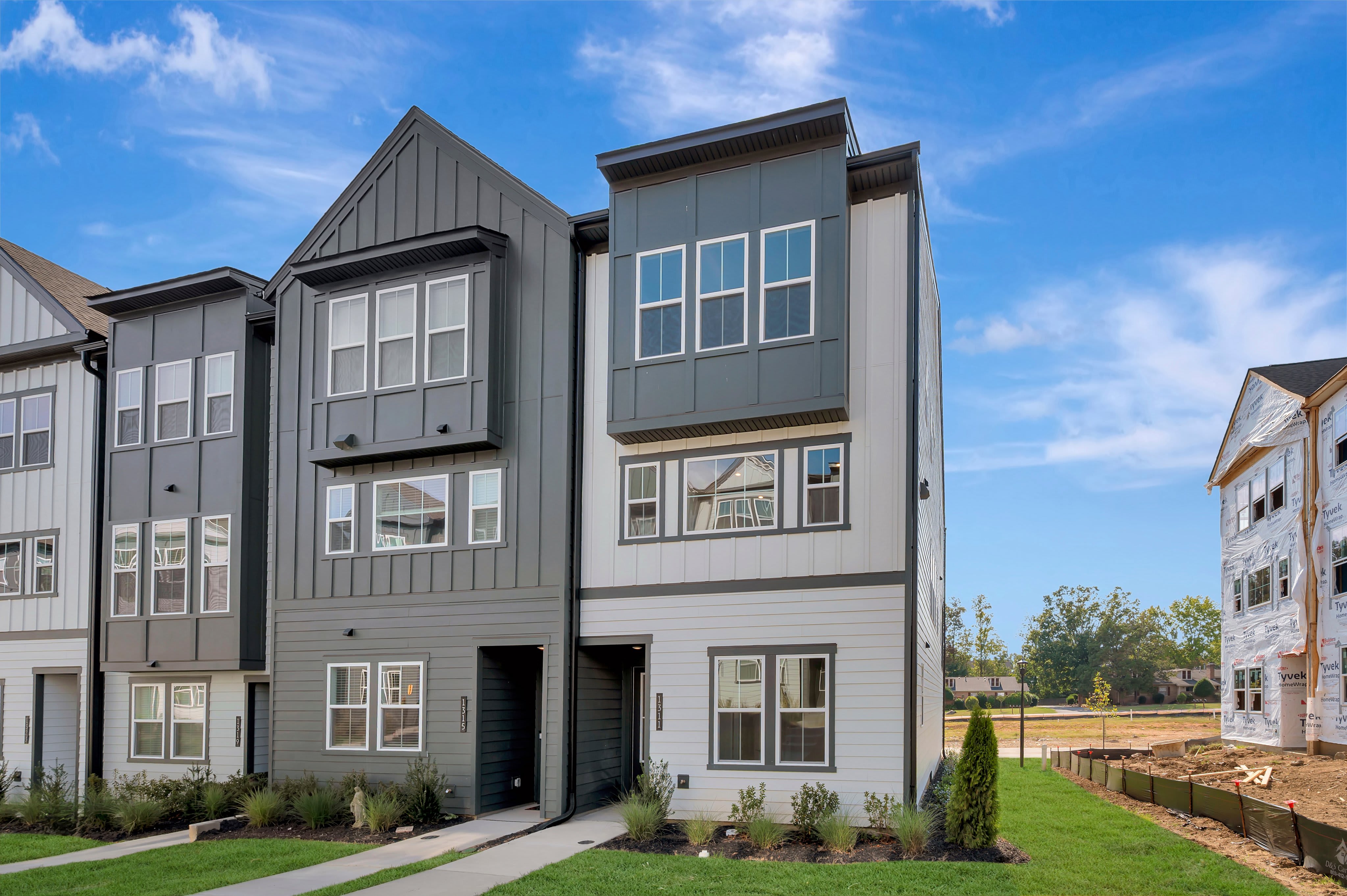 Modern multi-story townhomes with grey and white siding stand in the foreground against a bright blue sky with wispy clouds and a construction site in the background.