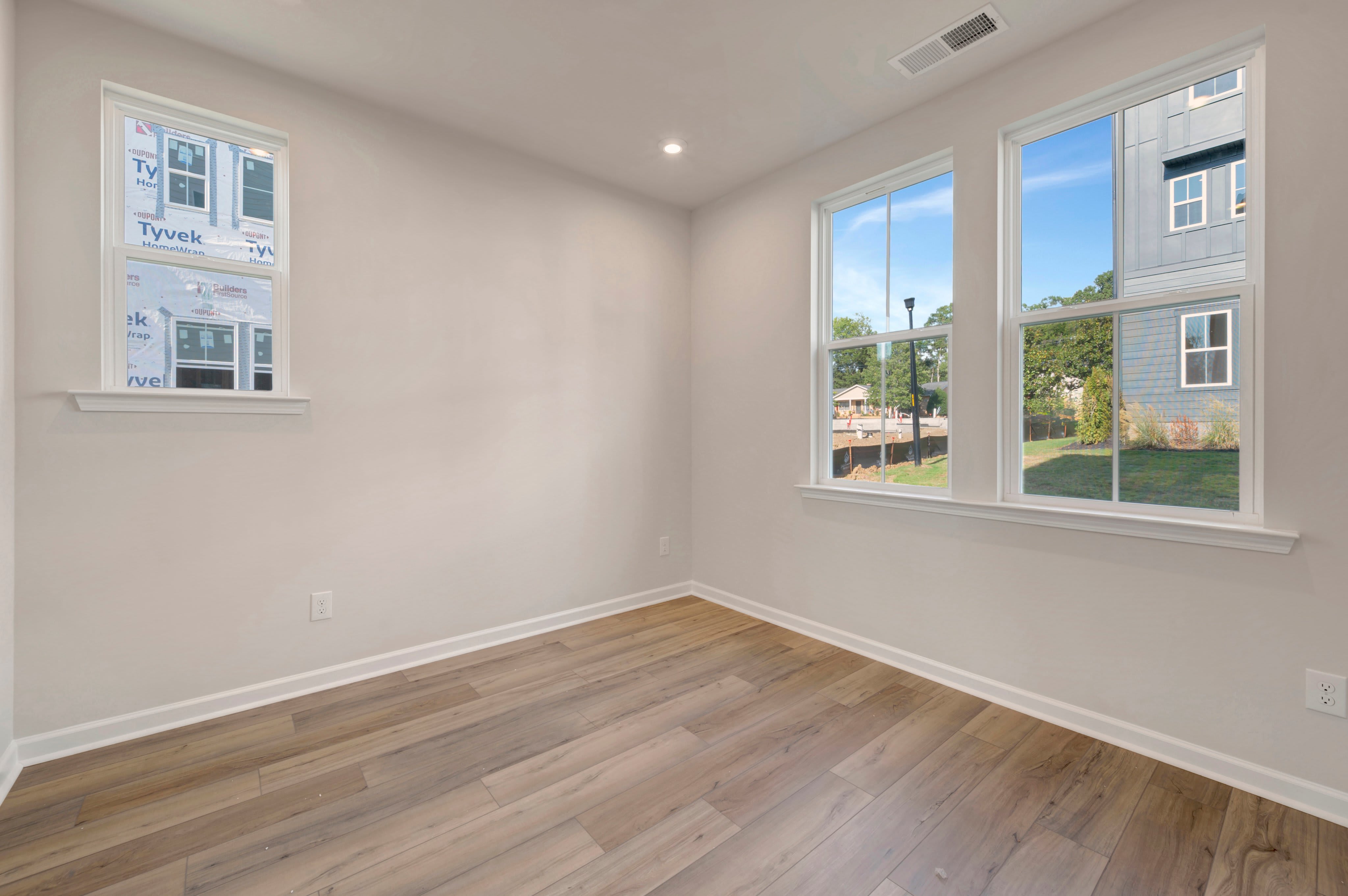 An empty room with light wood flooring and white walls features a small window on the left and two larger windows on the right overlooking a residential yard.