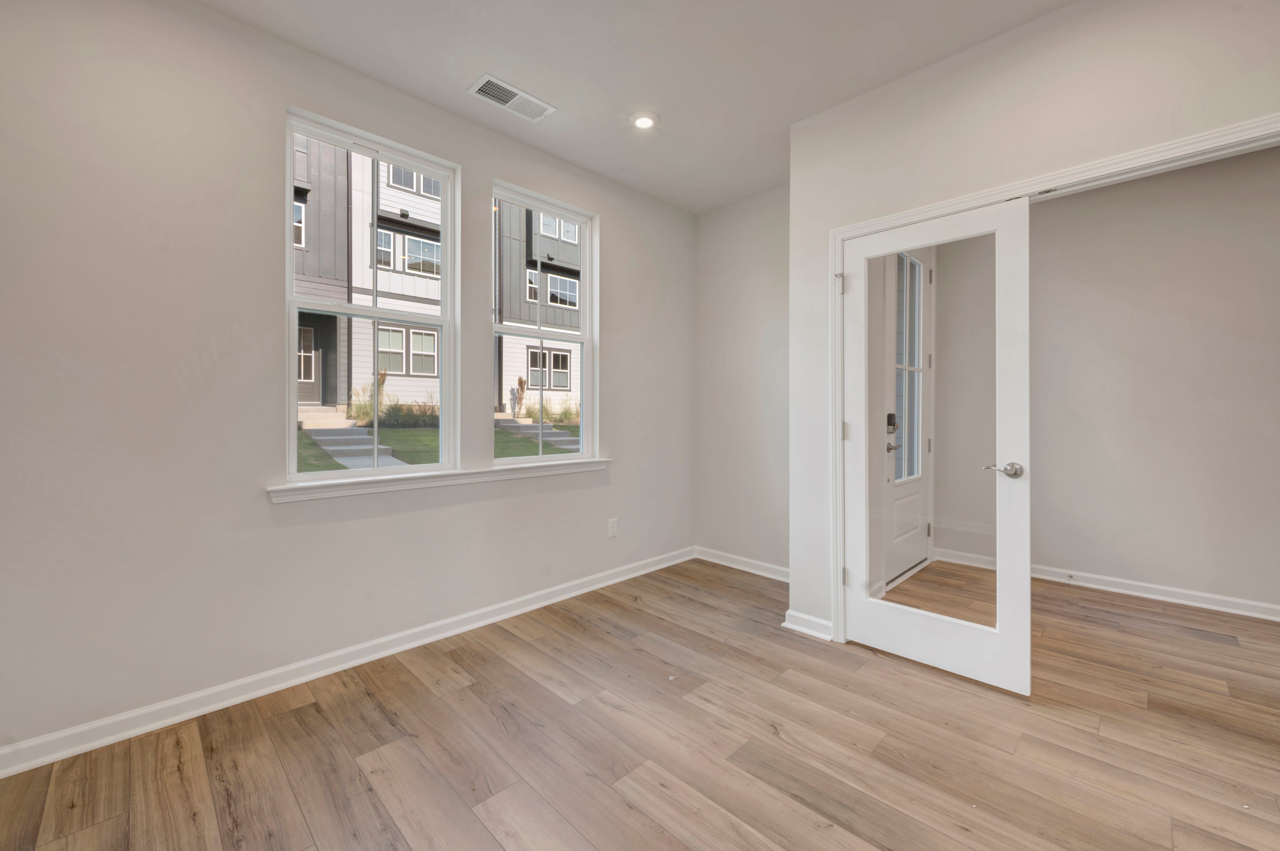 An empty room with light wood flooring and white walls features two windows looking out at neighboring houses and an open white door leading to another space.