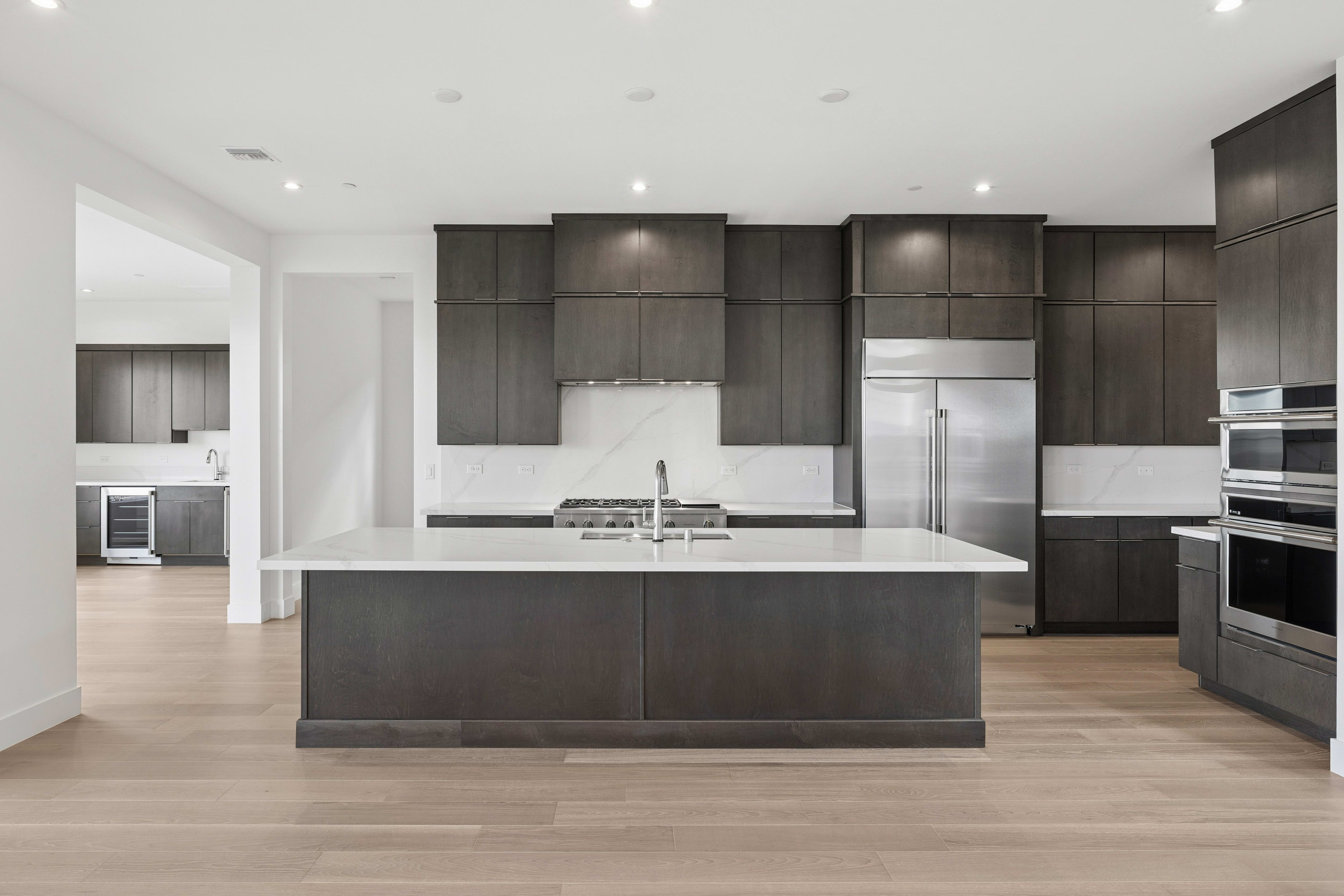 A large dark wood kitchen island with a white countertop sits in the foreground of a modern kitchen featuring dark cabinetry and stainless steel appliances in the background.