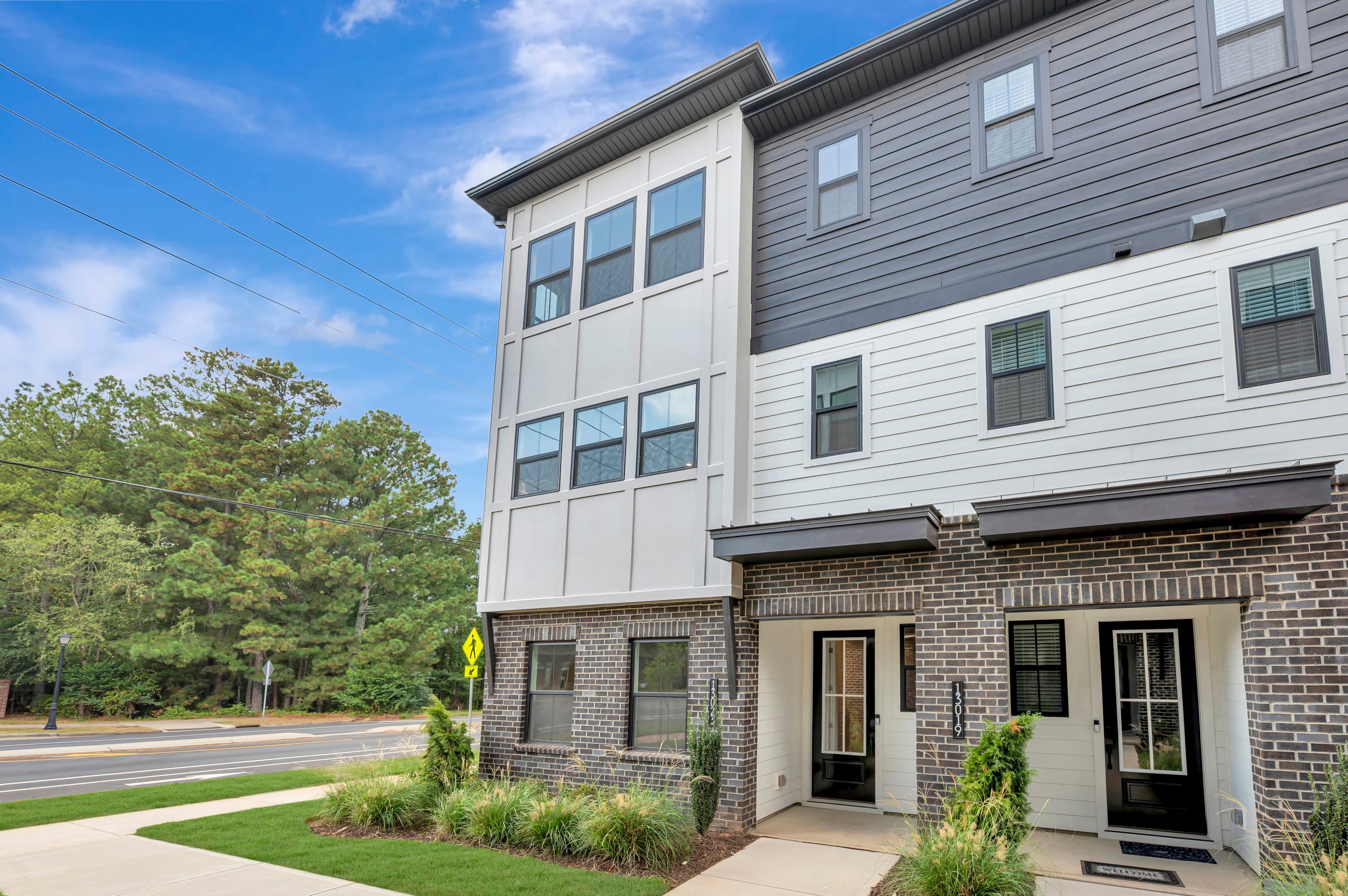 A modern multi-story townhouse with grey and white siding and brick accents stands before a backdrop of lush green trees and a bright blue sky.