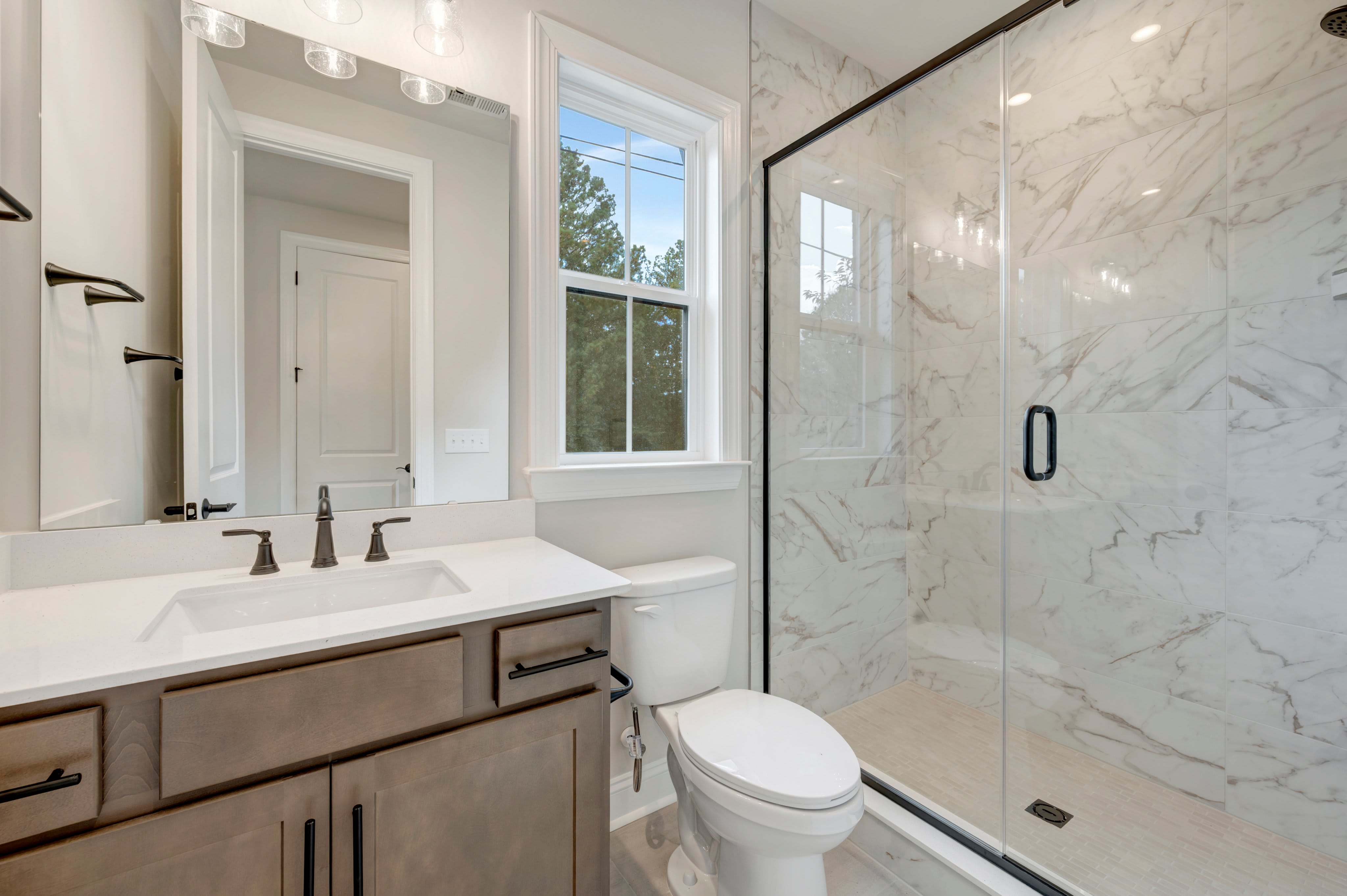 A modern bathroom features a wooden vanity with a white countertop in the foreground and a white toilet next to a glass-enclosed marble shower in the background.