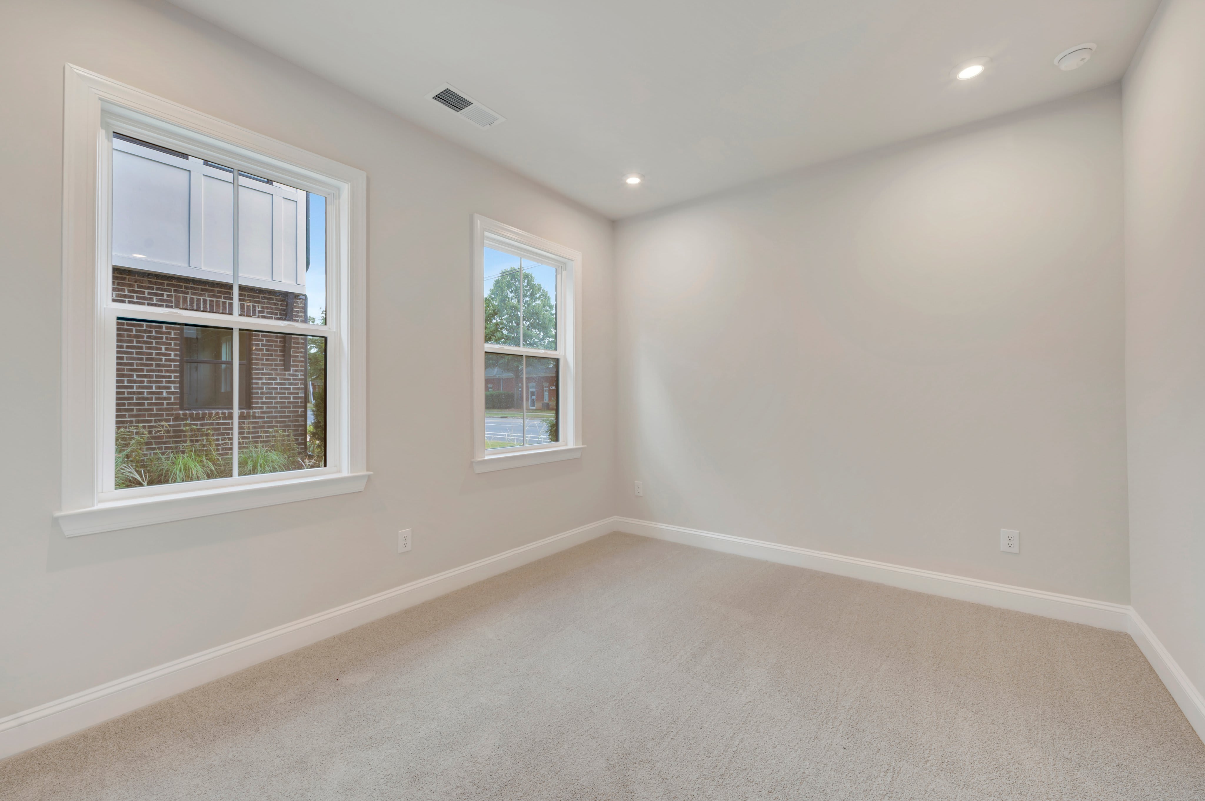 An empty room with beige carpeting and white walls features two windows looking out onto a brick building and greenery.