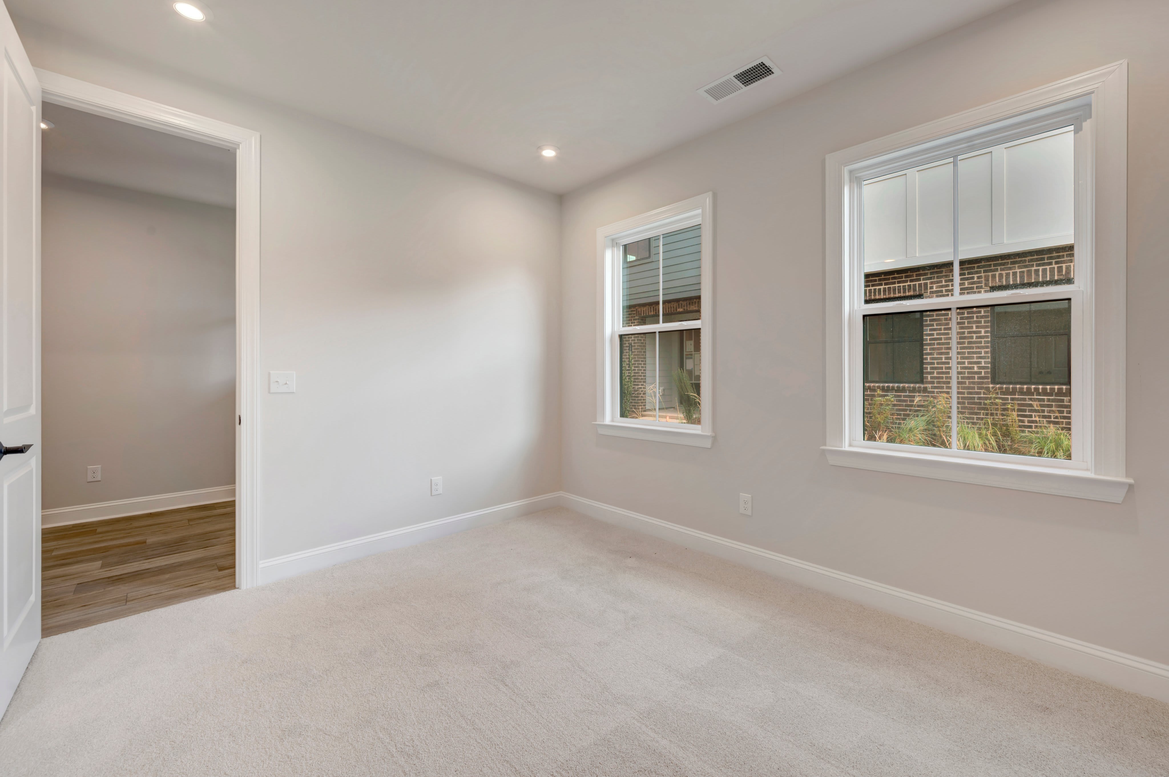 An empty room with light gray walls and beige carpeting features two windows looking out onto a brick building and an open doorway leading to another room with hardwood floors.