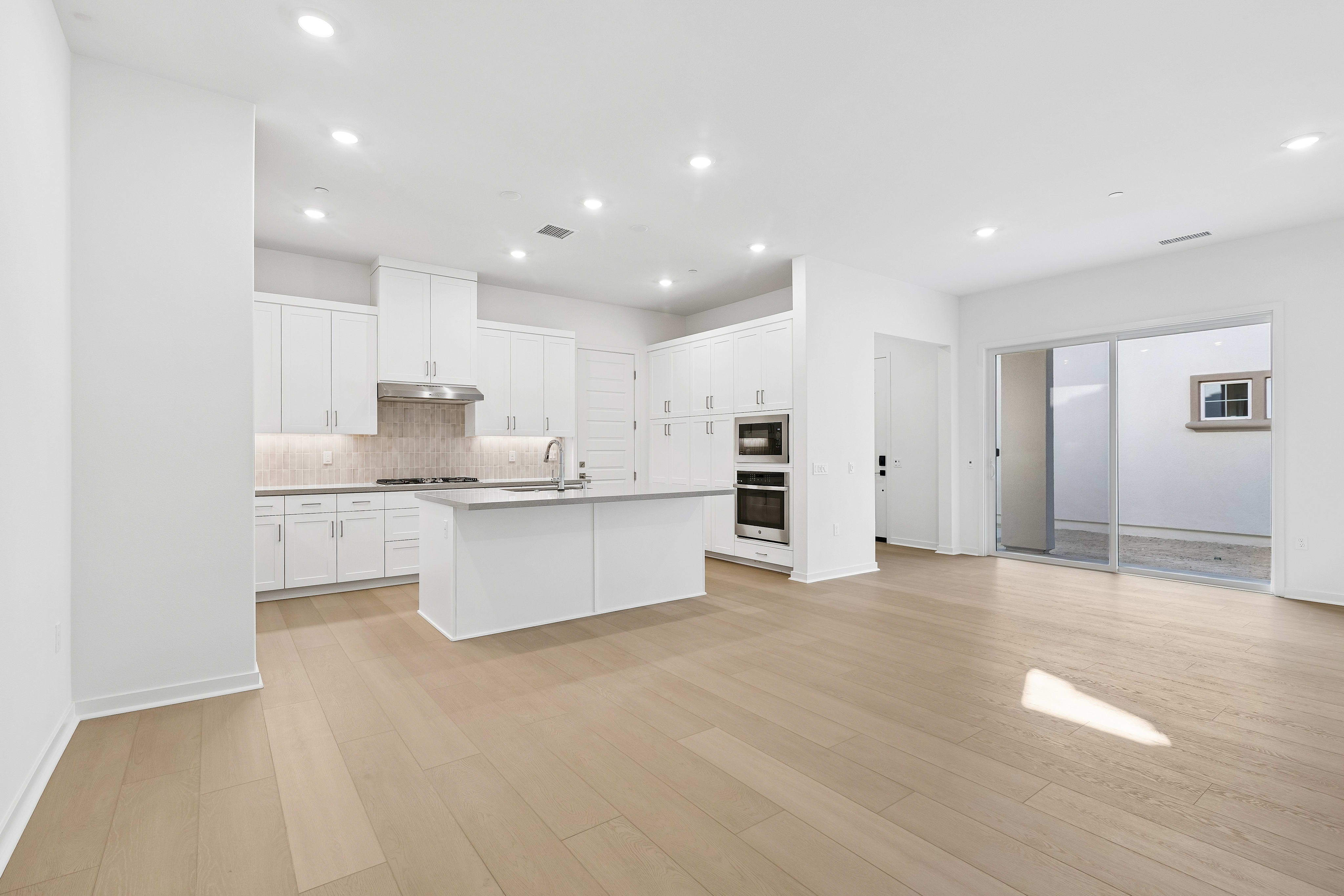 A spacious open-concept room features light wood flooring in the foreground and a modern white kitchen with a central island in the background.