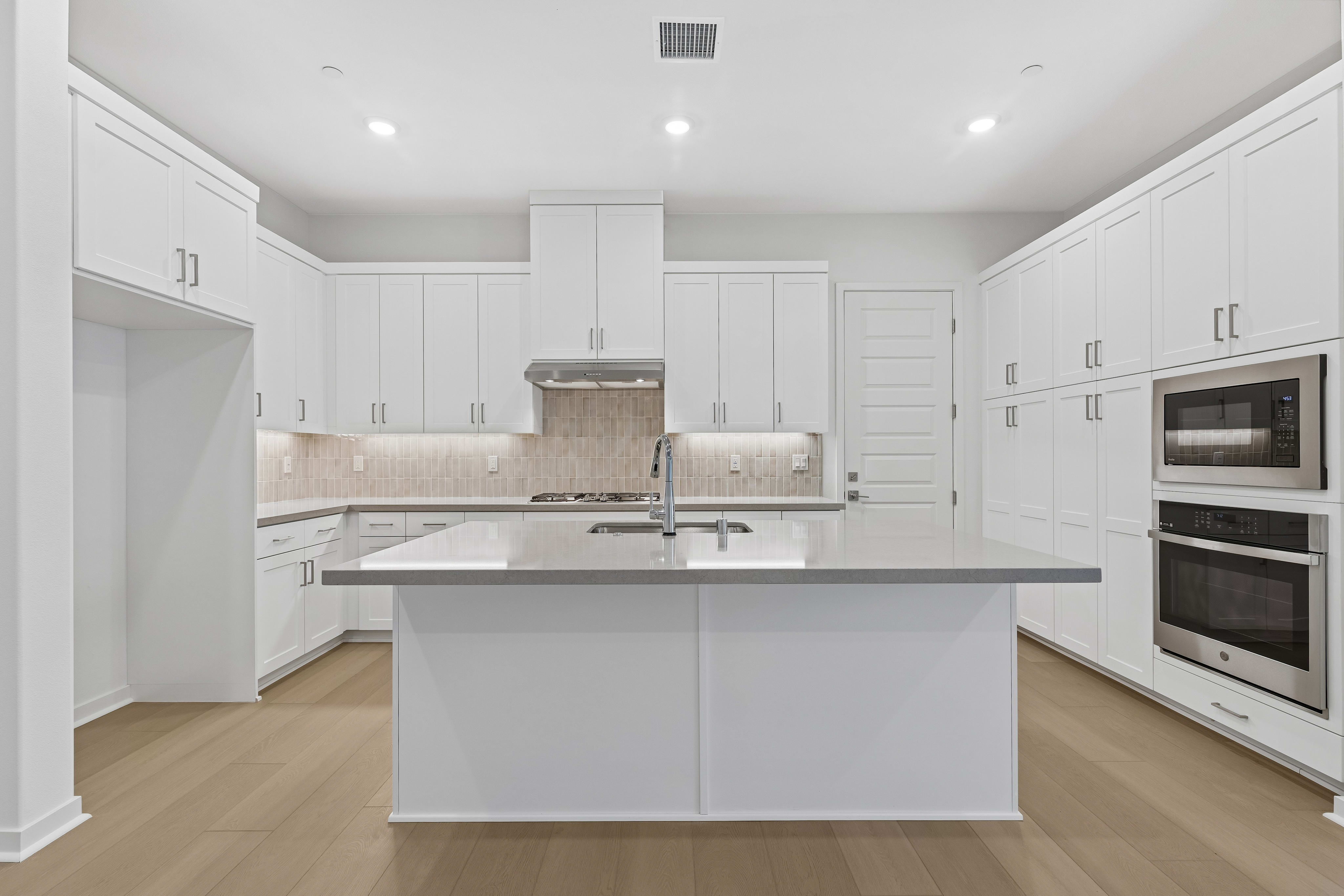 A white kitchen island with a grey countertop sits in the foreground of a bright room featuring white cabinetry and light wood flooring.