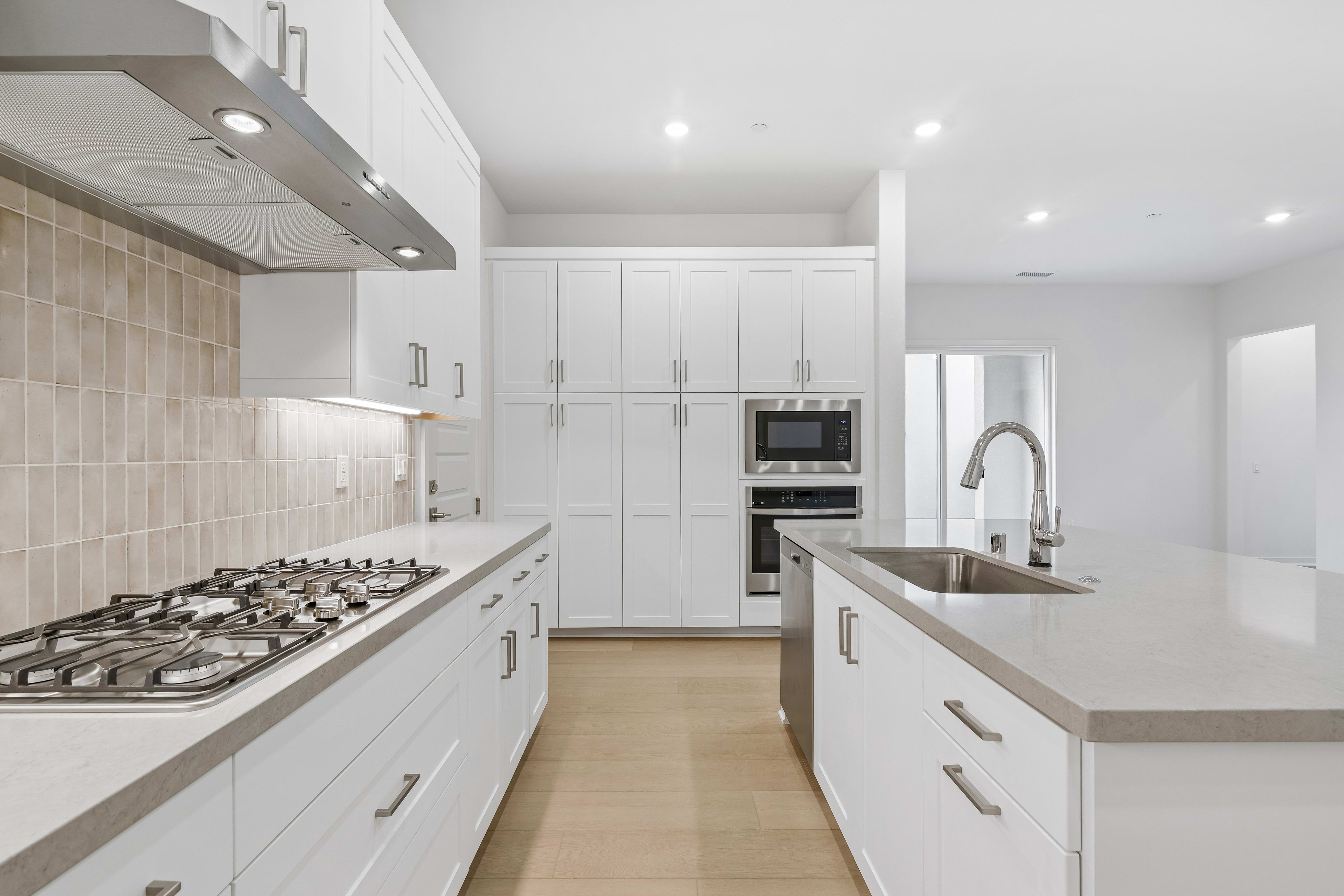 A modern white kitchen features a gas stovetop and range hood in the foreground, with a large island and built-in wall ovens in the background.