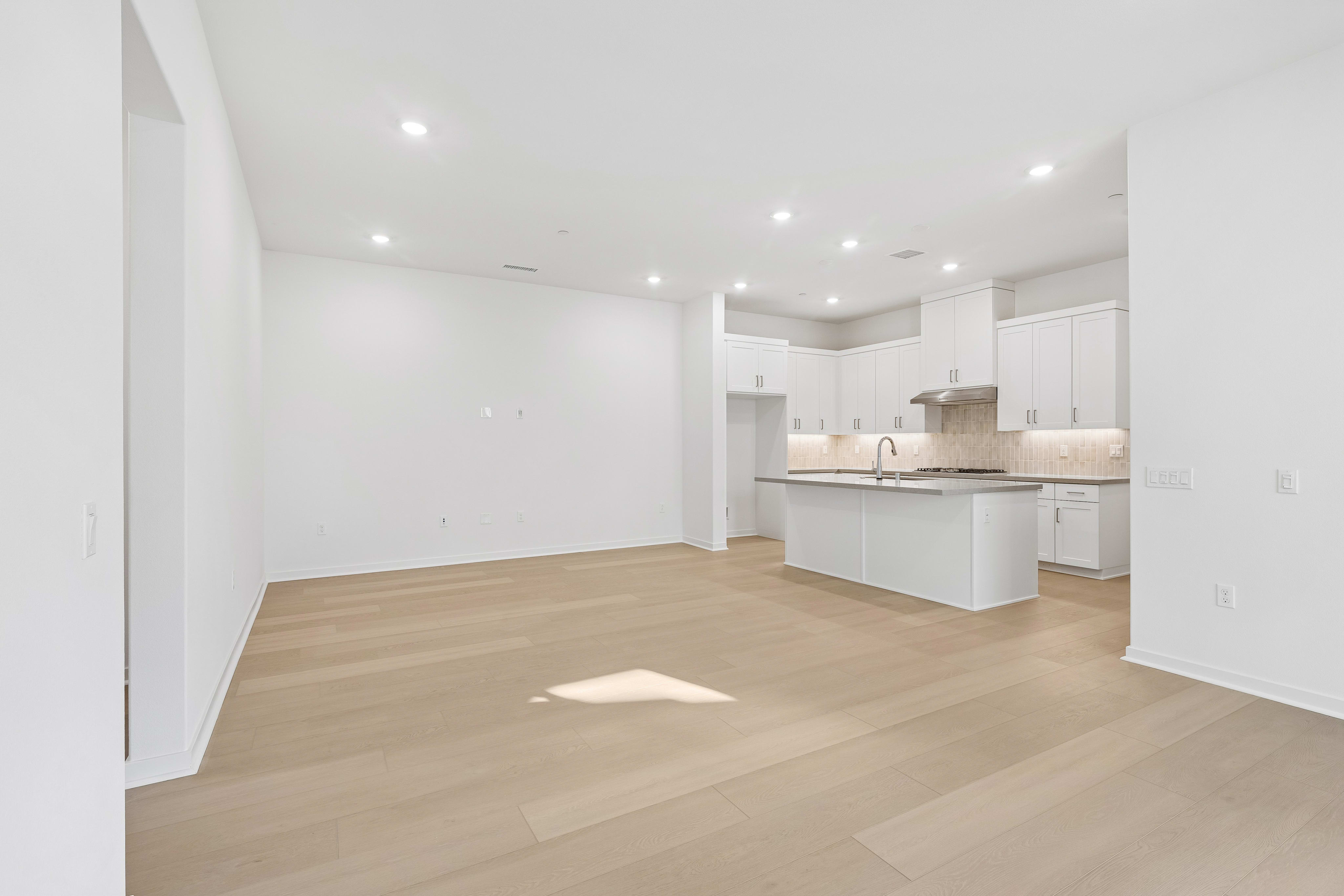 An empty room with light wood flooring leads to a modern white kitchen in the background.