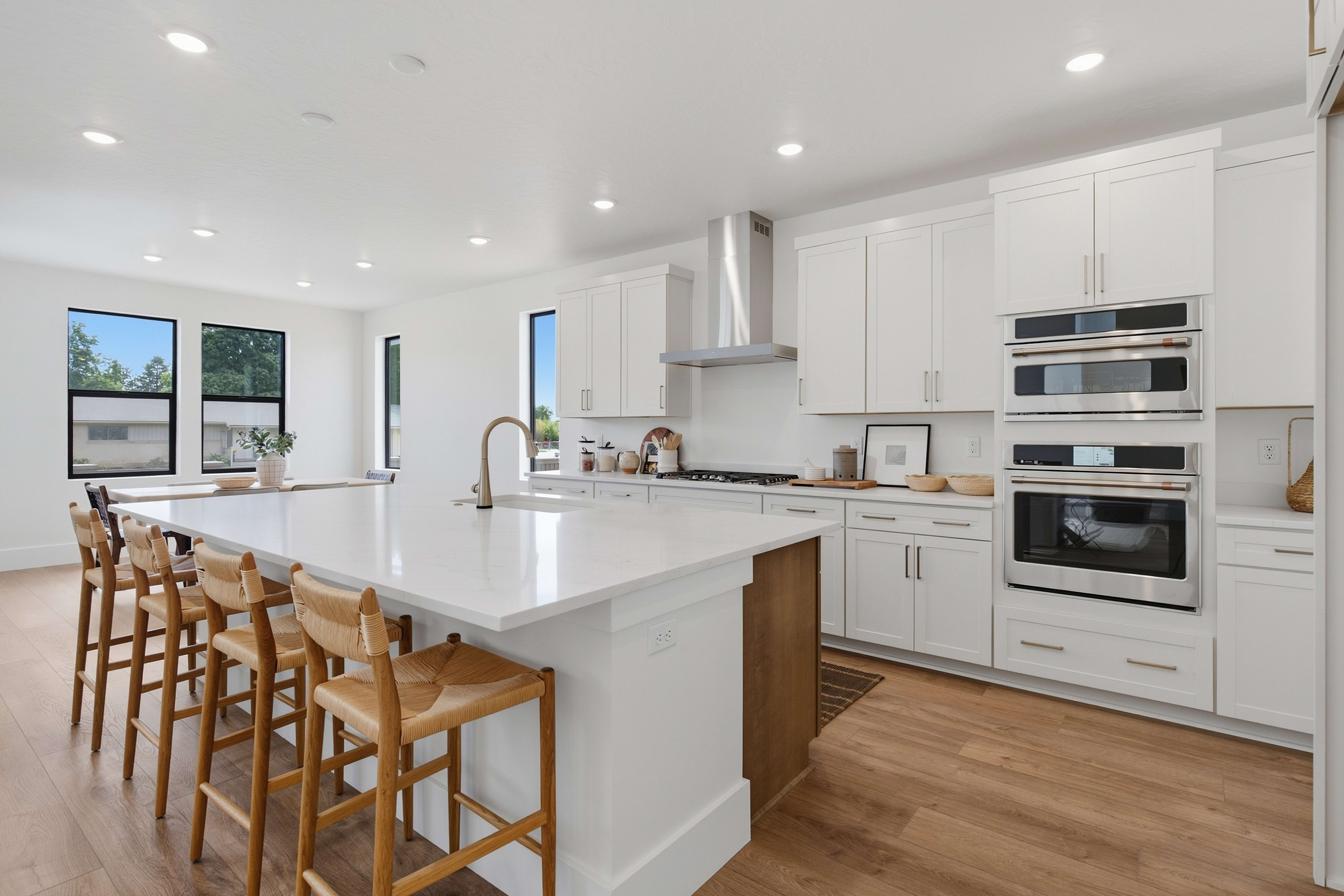A white kitchen island with wooden barstools sits in the foreground of a bright room featuring white cabinetry and stainless steel appliances in the background.