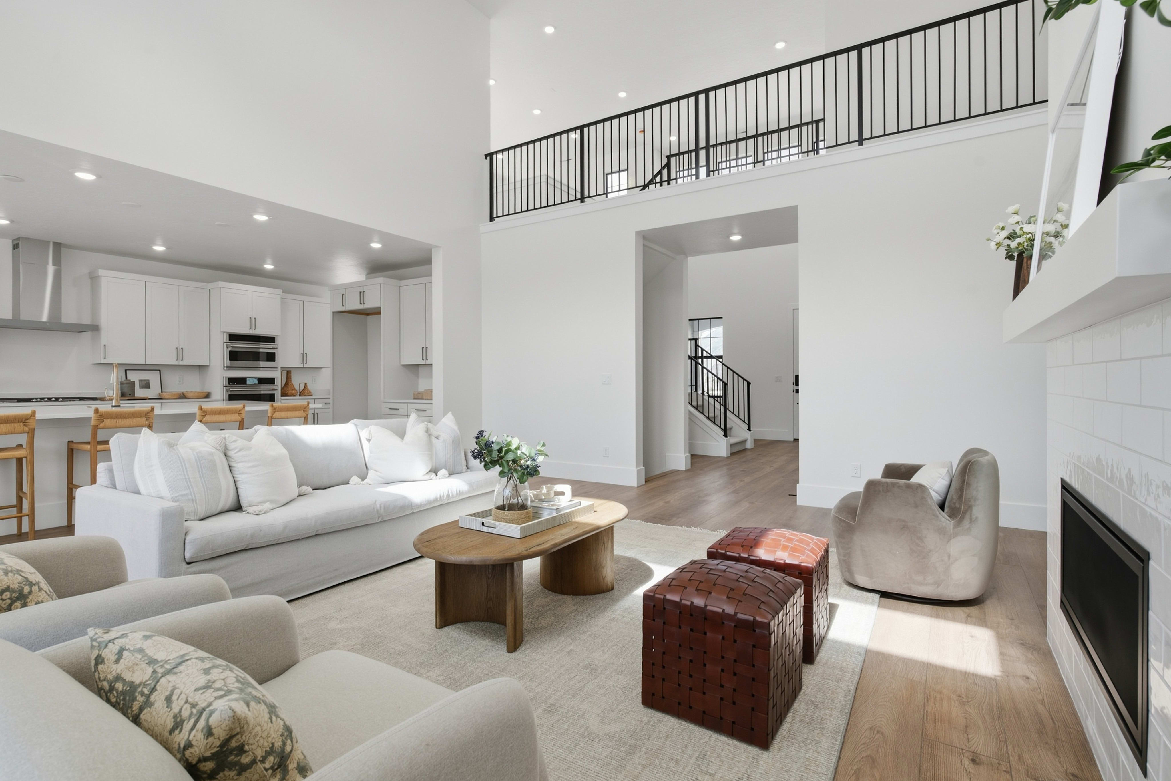A modern white living room with a plush sofa and wooden coffee table sits in the foreground, while a kitchen and an upper-level balcony with black railings are visible in the background.