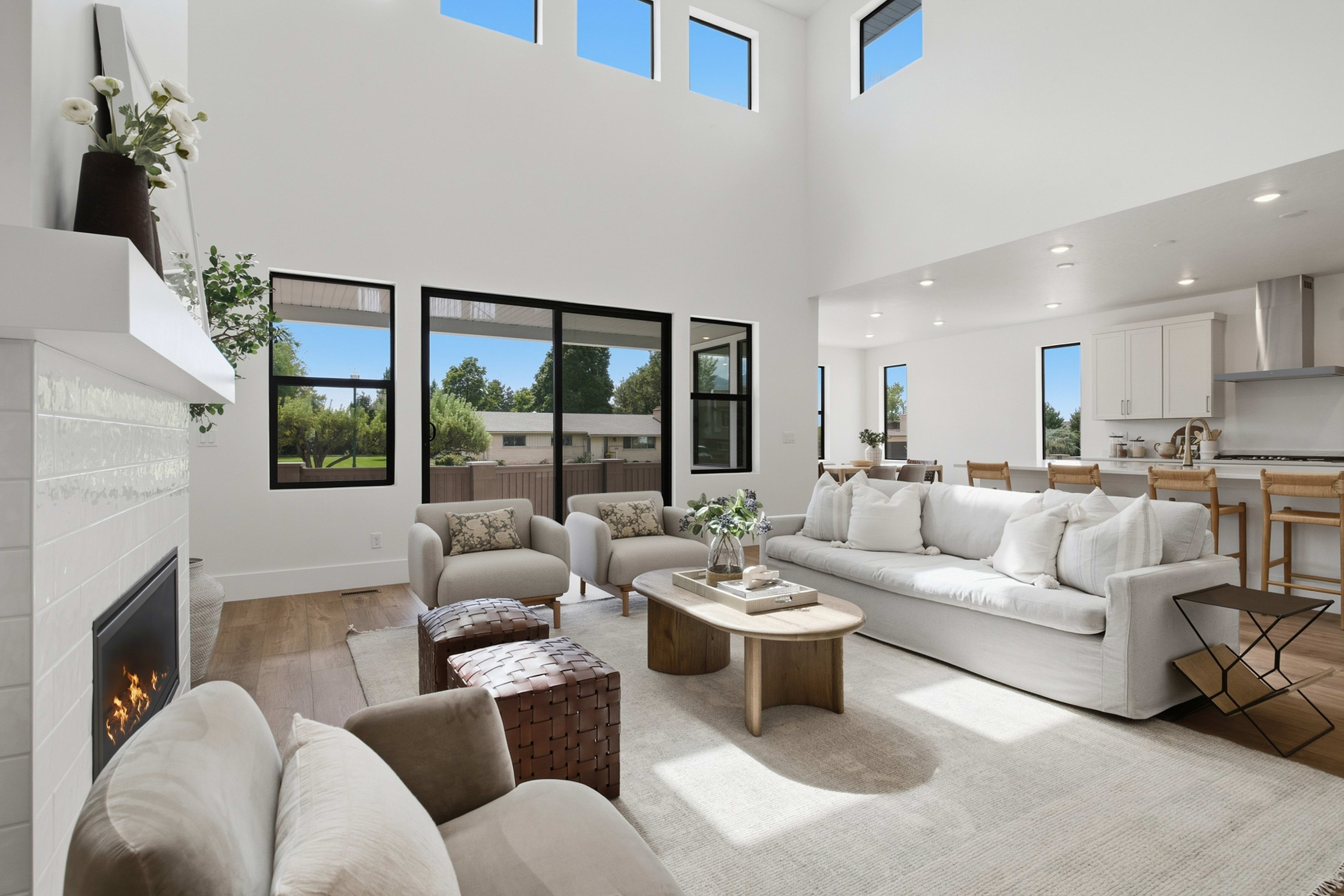 A modern living room with a white sofa and fireplace in the foreground opens up to a bright, high-ceilinged space and a kitchen in the background.