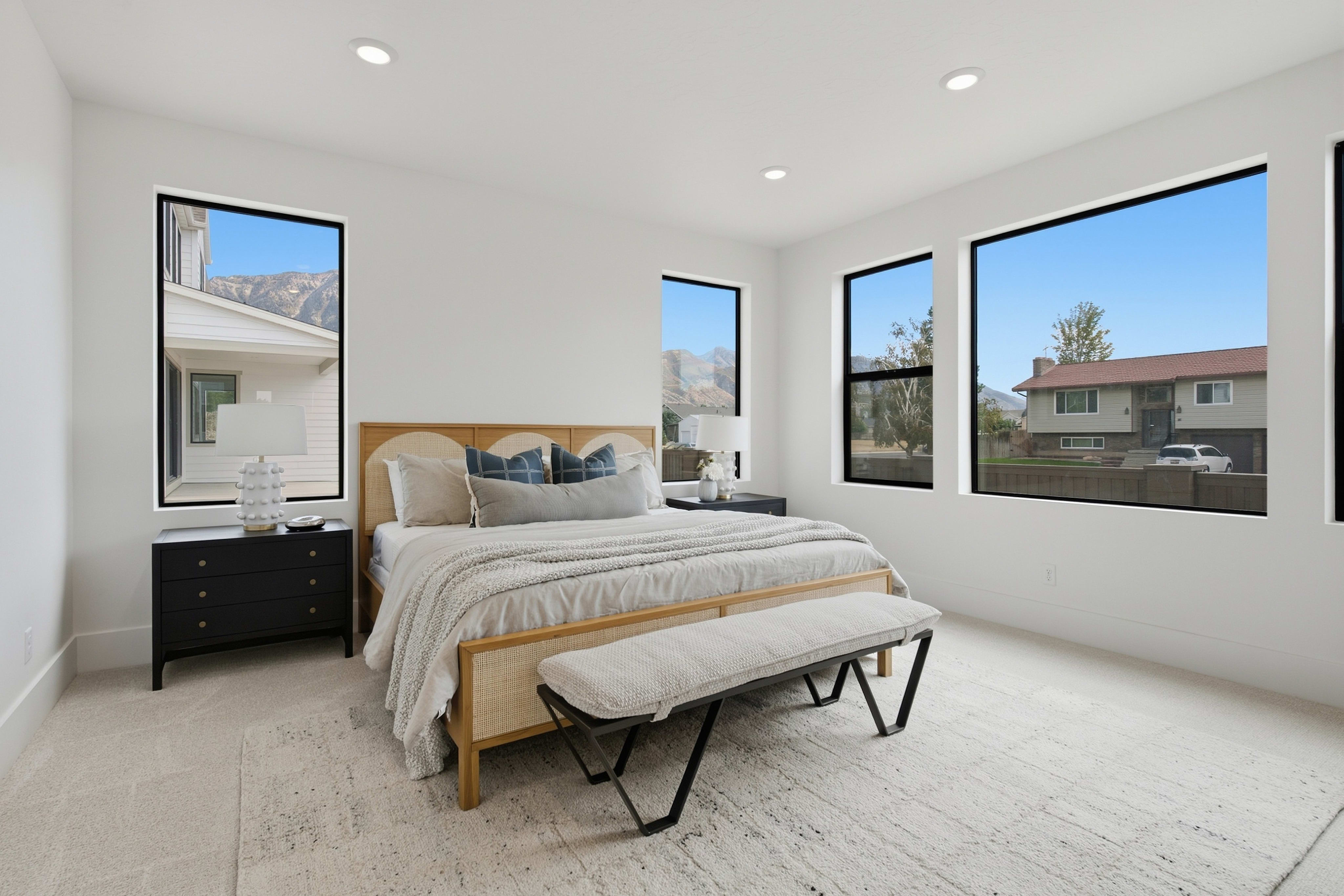 A modern bedroom with a bed and bench in the foreground is surrounded by white walls and large windows overlooking a suburban landscape and mountains in the background.