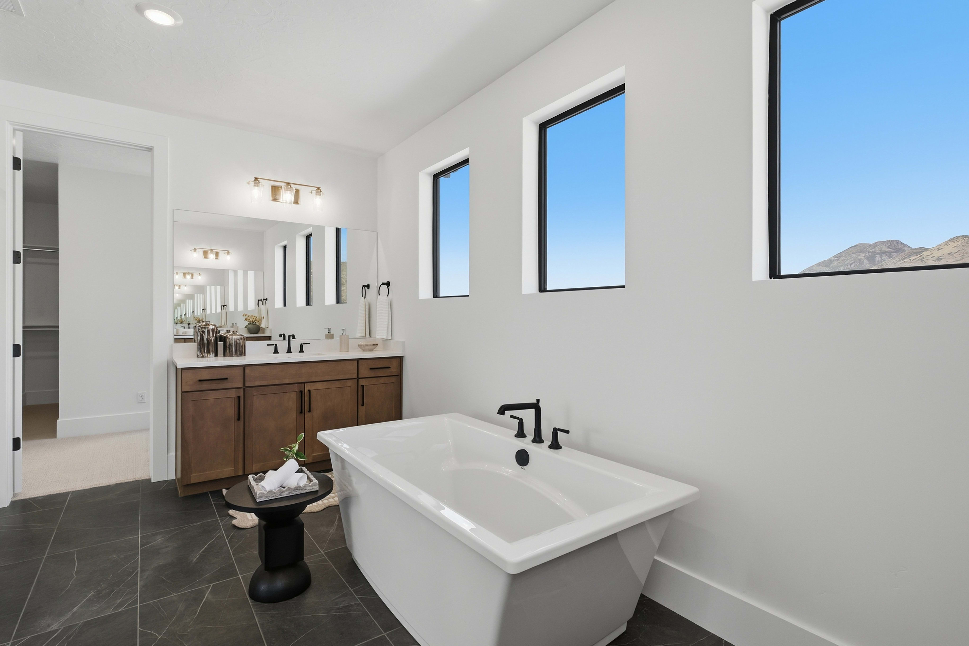 A modern white freestanding bathtub and small black side table sit in the foreground of a bright bathroom featuring a wooden vanity and windows overlooking a mountain landscape in the background.