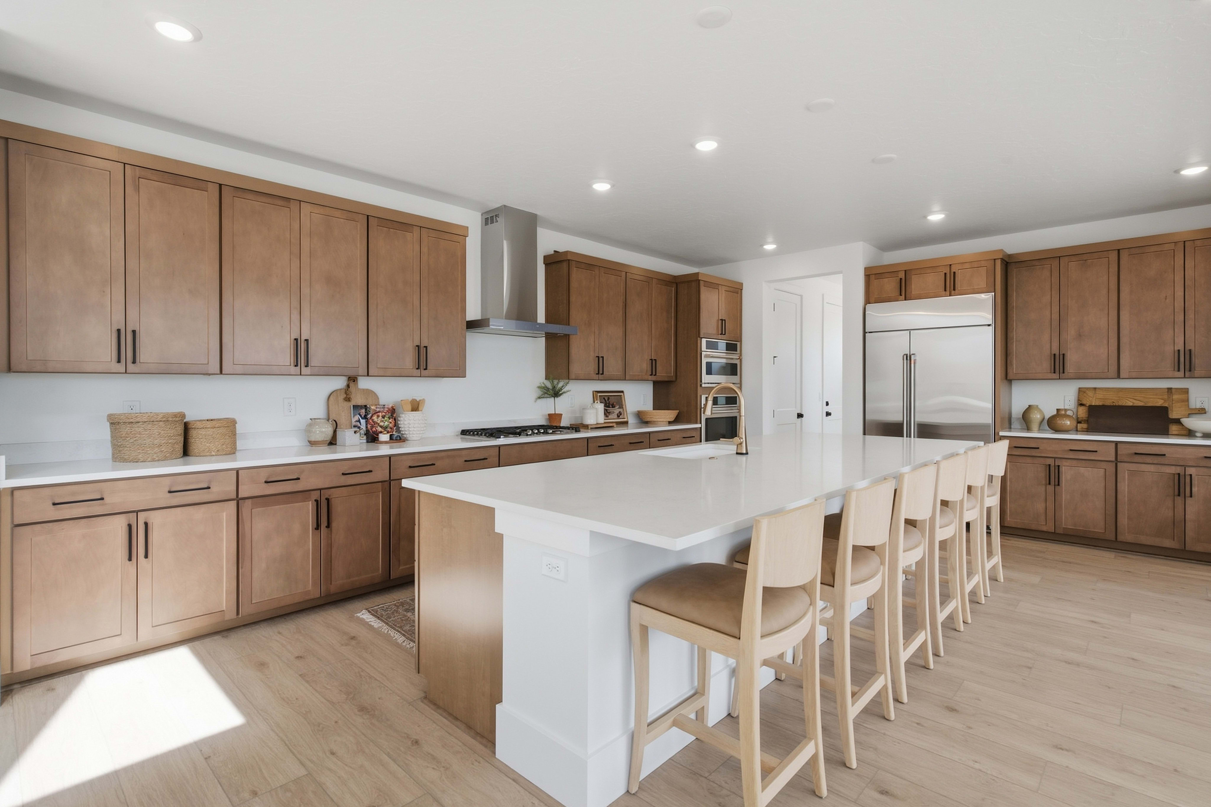 A long white kitchen island with wooden stools sits in the foreground of a modern kitchen featuring light wood cabinetry and stainless steel appliances in the background.