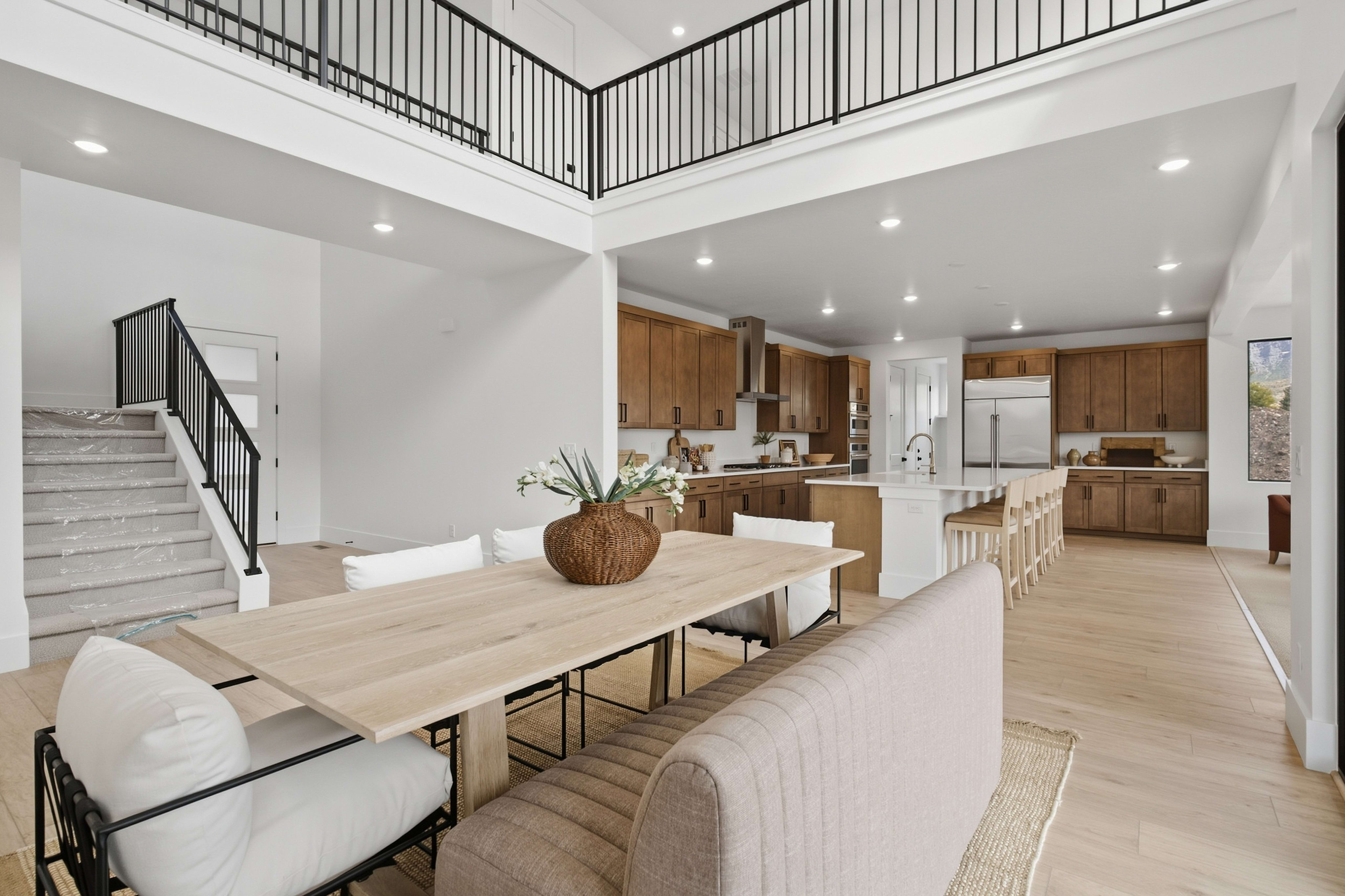 A light-colored wooden dining table and chairs sit in the foreground of a modern open-concept living space featuring a kitchen and an overlooking second-floor balcony in the background.