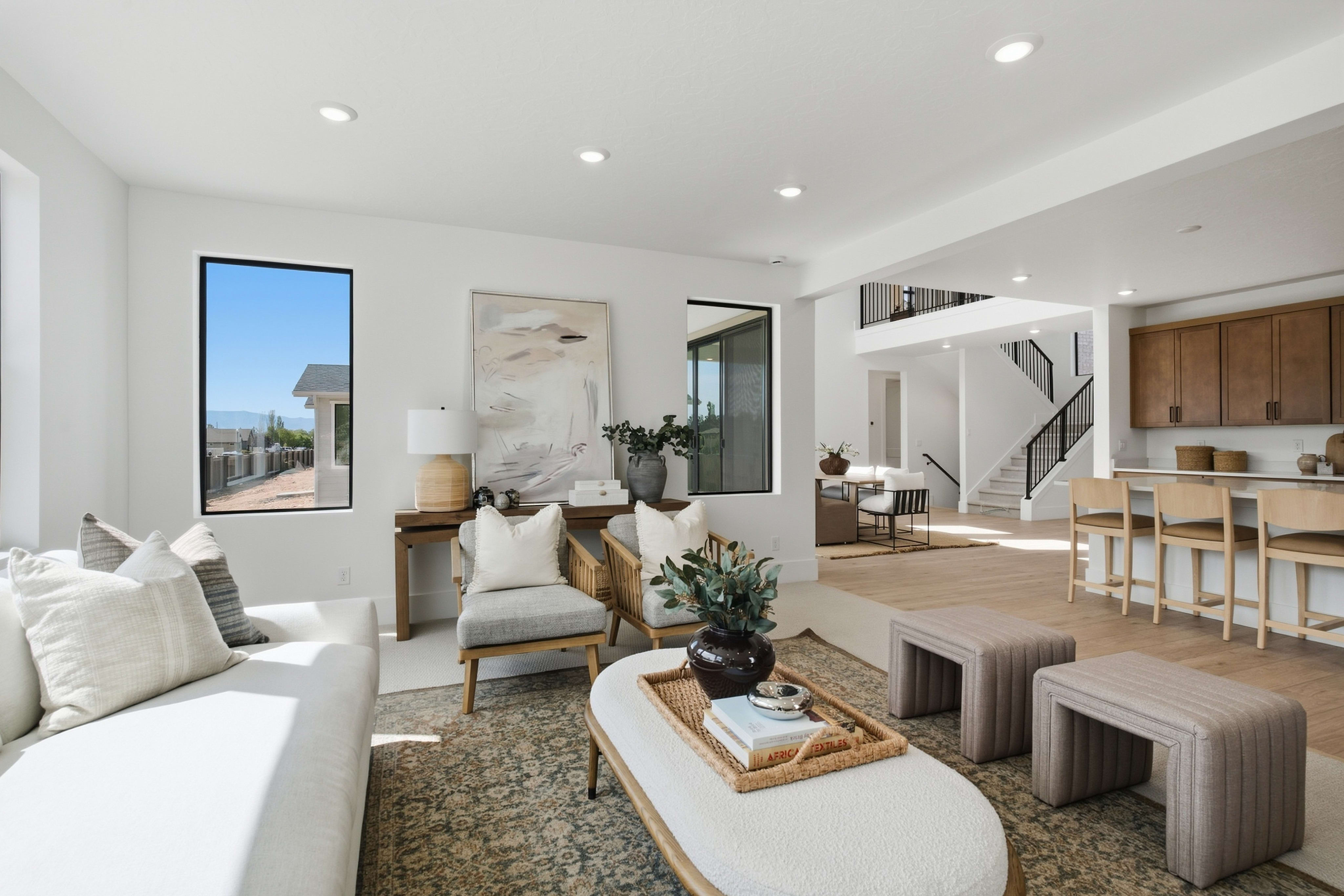 A bright, modern living room with a white sofa and textured ottomans in the foreground opens up to a dining area and staircase in the background.